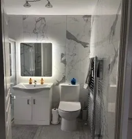 Bathroom with a toilet, vanity cabinet with sink and illuminated mirror, wall-mounted heated towel rack, marble-patterned wall tiles and tiled floor.