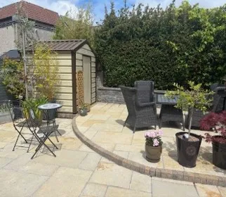 Outdoor paved patio with wicker chairs, a small metal bistro table and chairs, several potted plants, and a garden shed against a tall hedge.