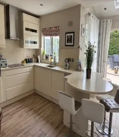 Kitchen with L-shaped cream cabinets, a round breakfast bar with two stools, a window with a patterned blind, and patio doors leading outside.