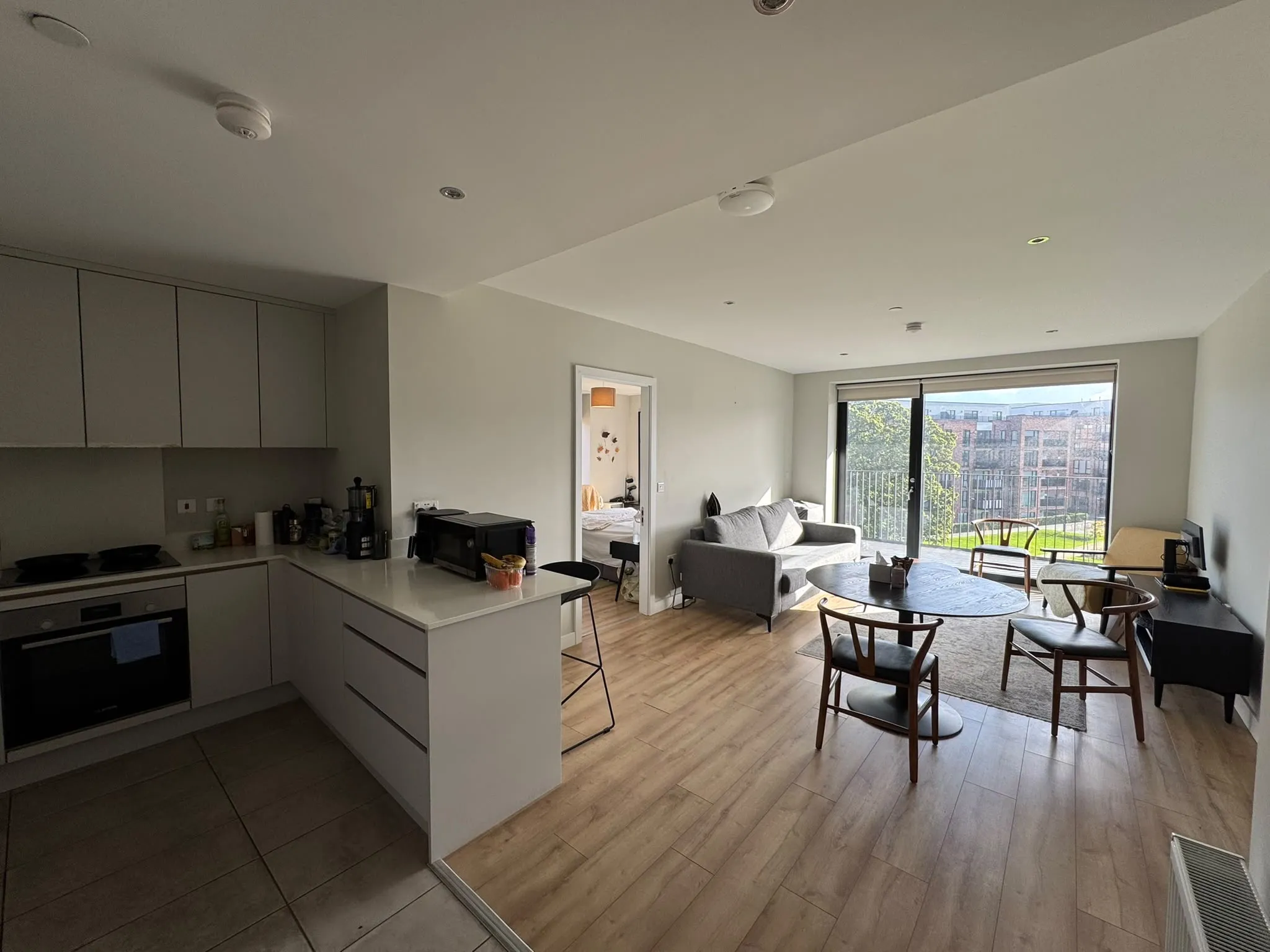 Open-plan living area showing a kitchen island with appliances, a sofa, round dining table with chairs, and a sliding glass door to a balcony.
