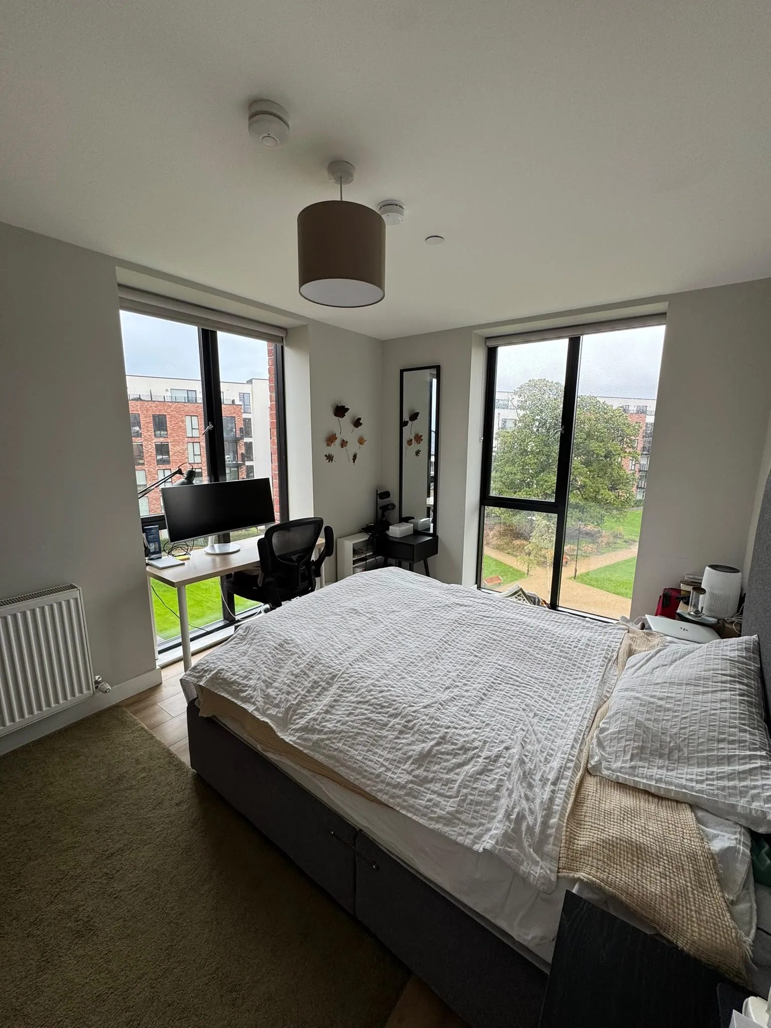 Bedroom with a double bed, bedside table, desk with monitor and chair near two large windows, and a ceiling pendant light.