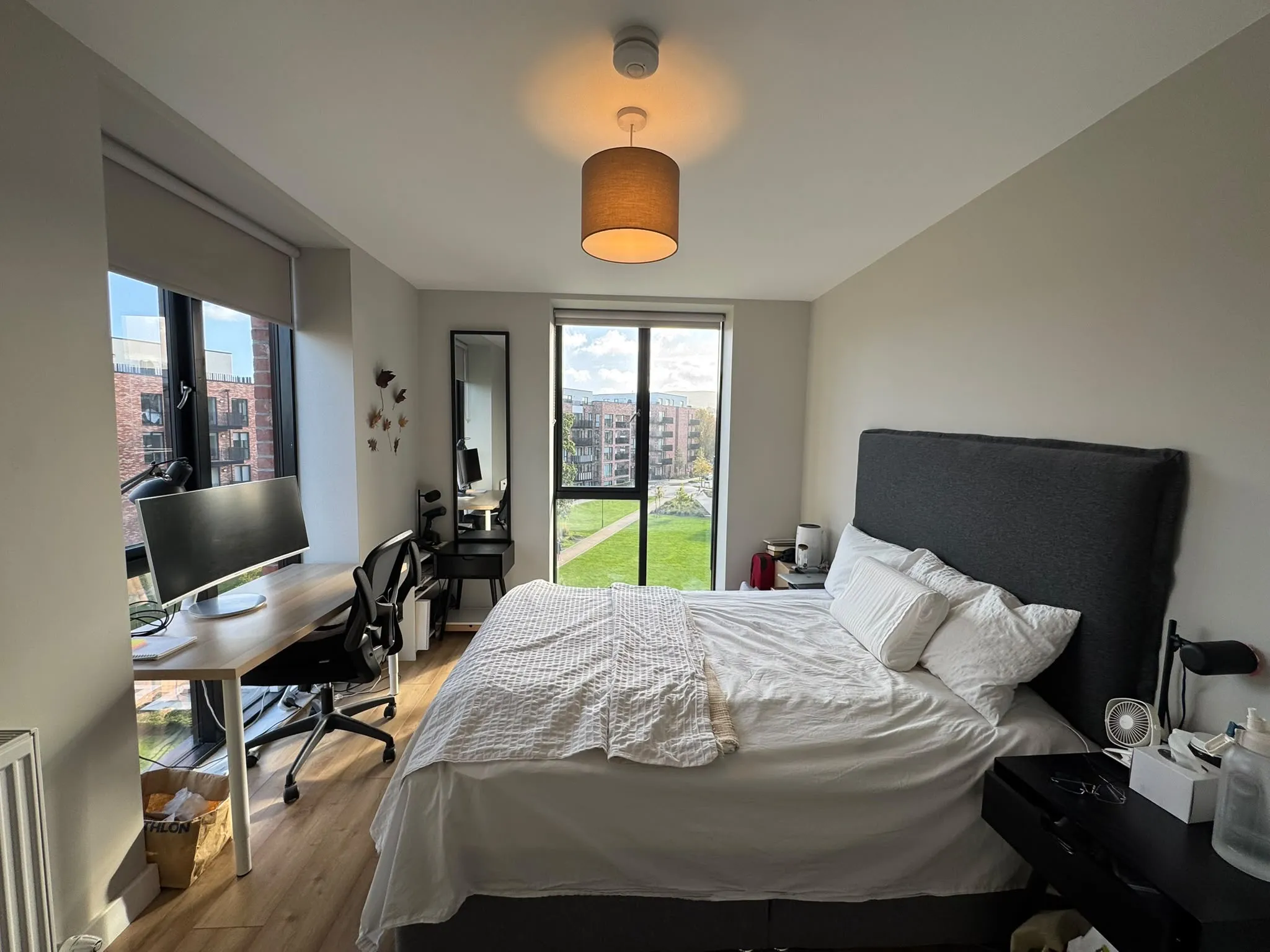 Bedroom with a double bed and tall headboard, bedside table, desk with a monitor and office chair, and a large window with a view