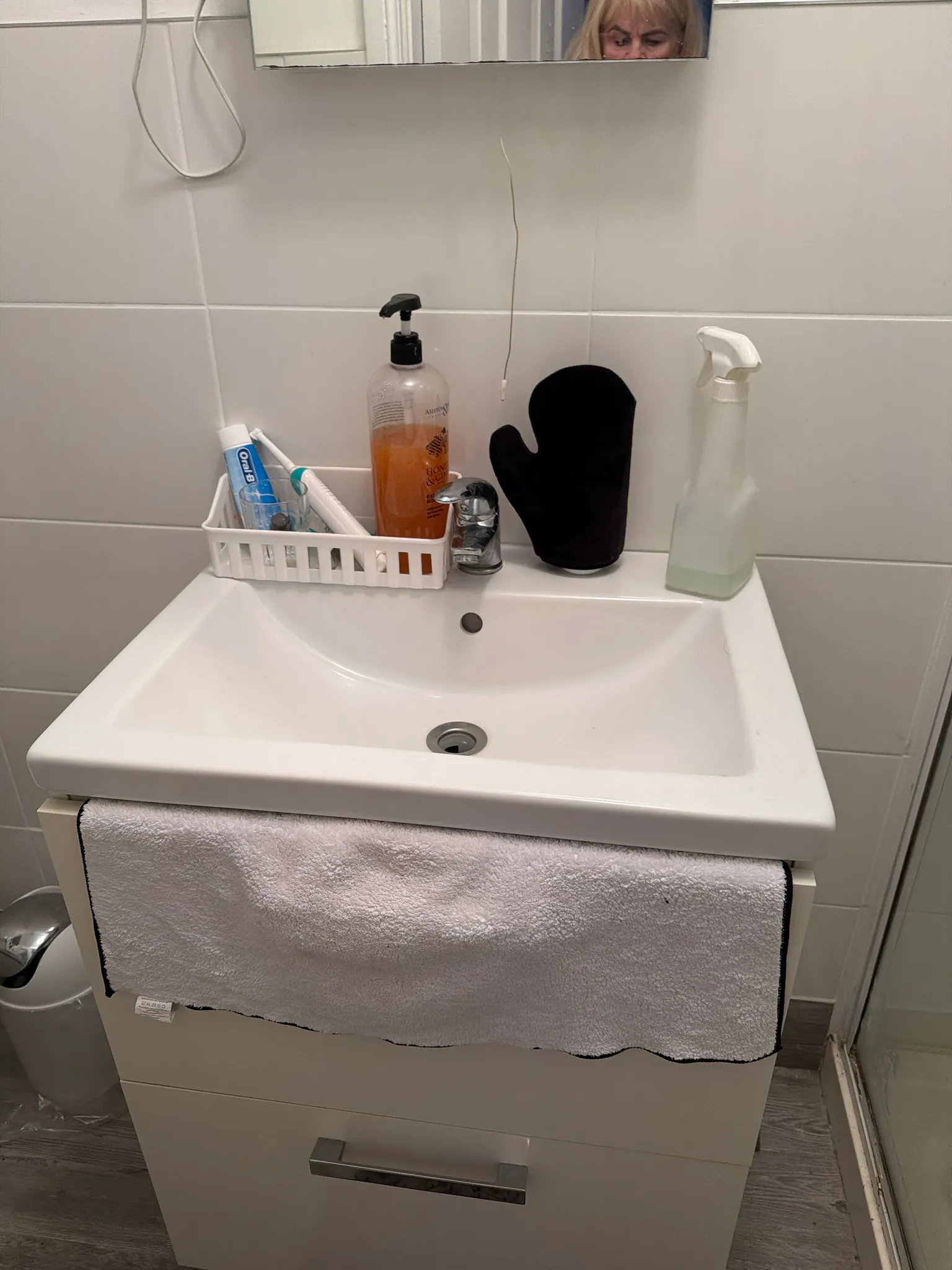 Bathroom sink and cabinet with faucet, liquid soap dispenser, toothbrush holder with toothpaste and brush, spray bottle, black mitt on the sink ledge, and a towel hanging on the cabinet; partial reflection visible in the