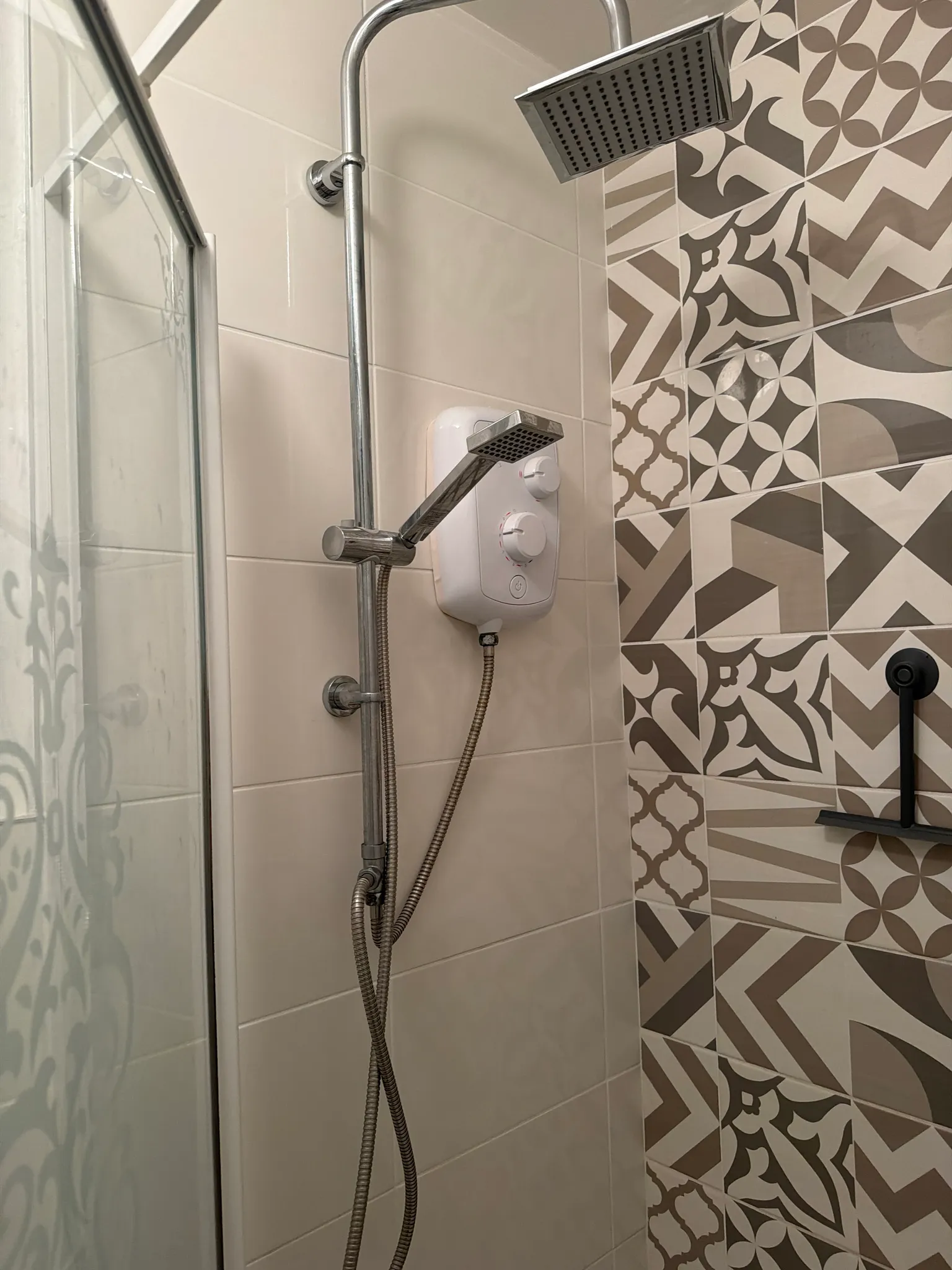 Bathroom shower area with wall-mounted electric shower unit, overhead square showerhead, handheld shower and patterned wall tiles.