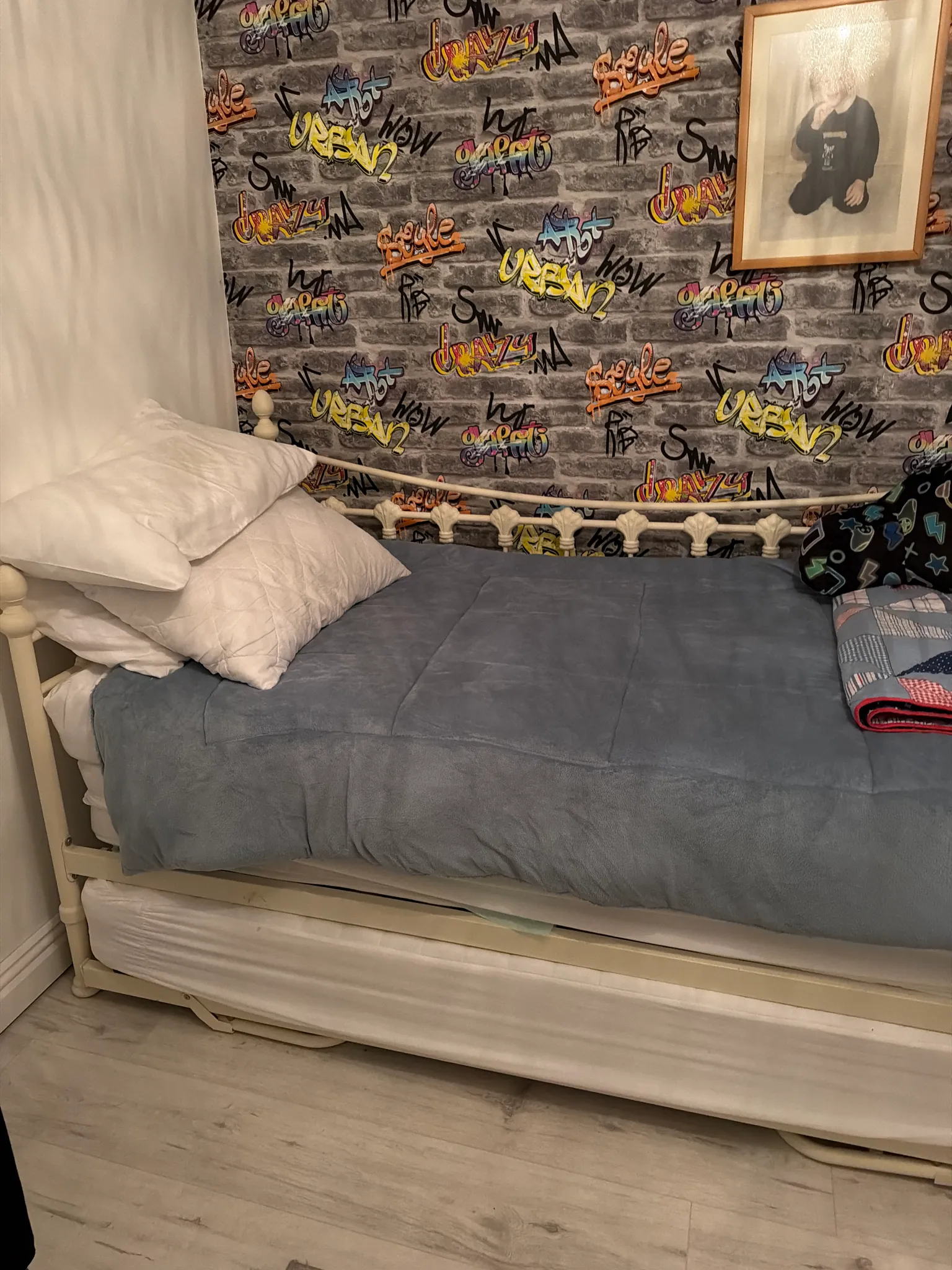 Bedroom with a single bed and pull-out trundle, pillows and blue blanket, graffiti-patterned wallpaper and a framed picture on the wall.
