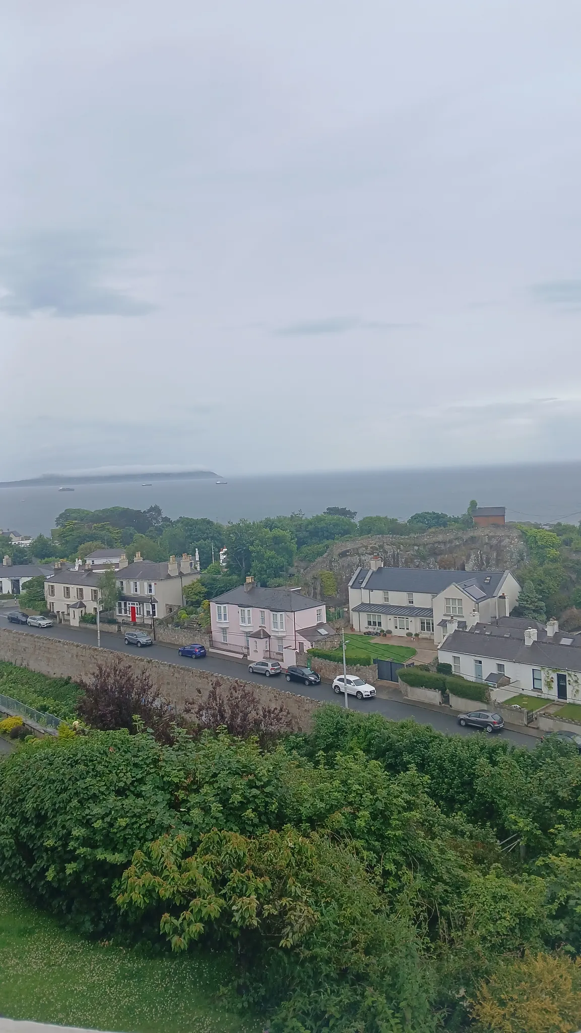 Exterior view from a higher level showing residential houses and a street with parked cars, green shrubs in the foreground and the sea with a distant shoreline under a cloudy sky.