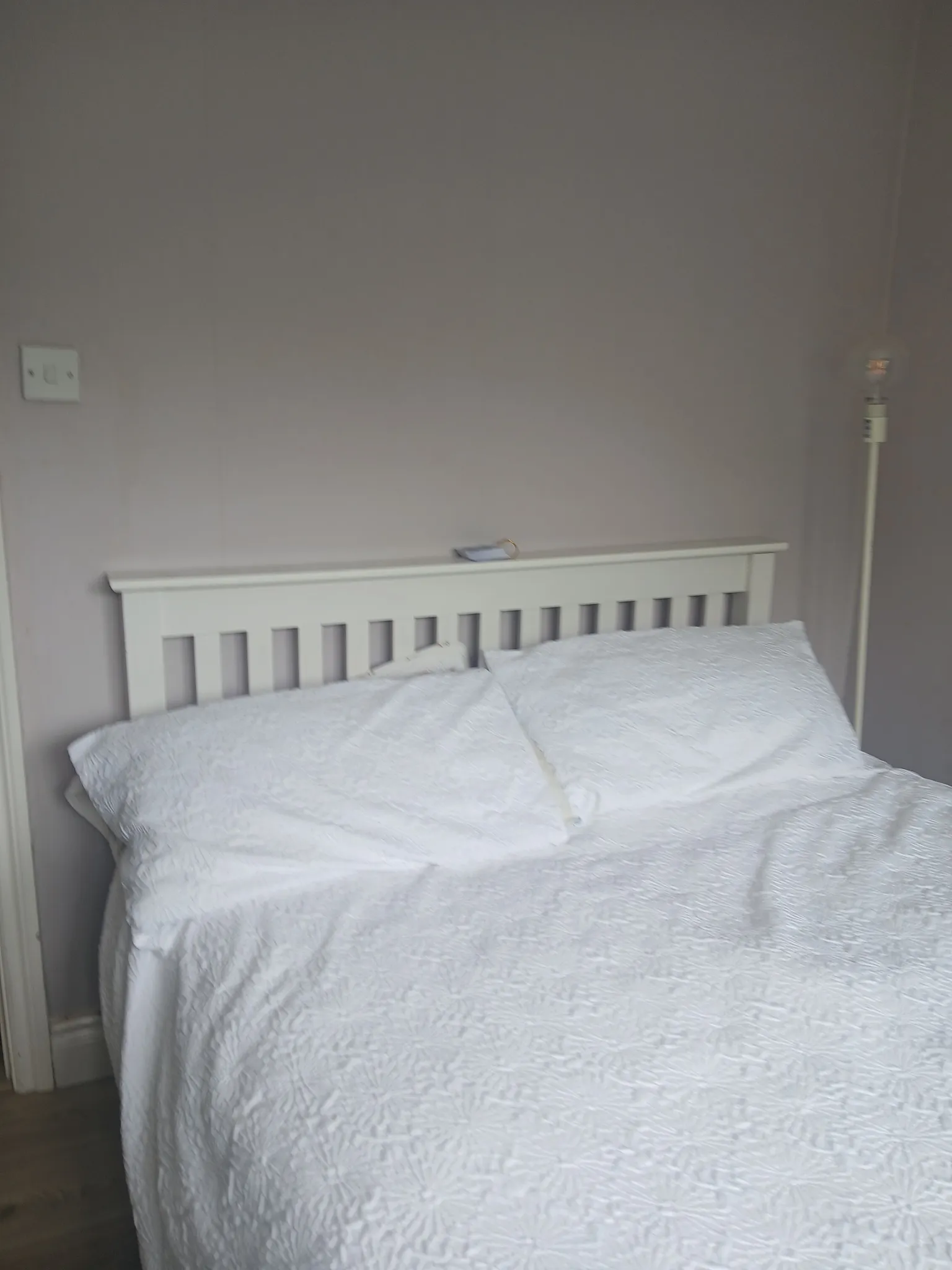 Bedroom with white bed and pillows, white slatted headboard, floor lamp to the right, and wall switch to the left.