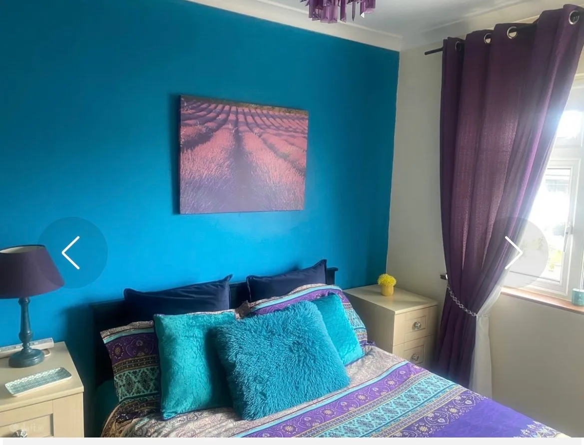 Bedroom with a double bed, teal accent wall, patterned bedding and teal pillows, nightstands with a lamp and small decor, purple curtains and a window.