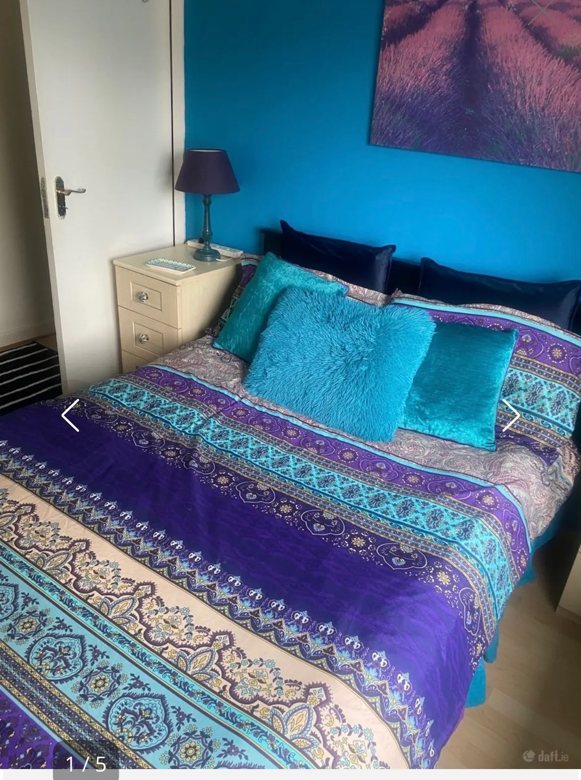 Bedroom with a double bed covered by a purple and turquoise patterned bedspread, several decorative pillows, a beige bedside drawer with a lamp, a blue painted wall and framed artwork.