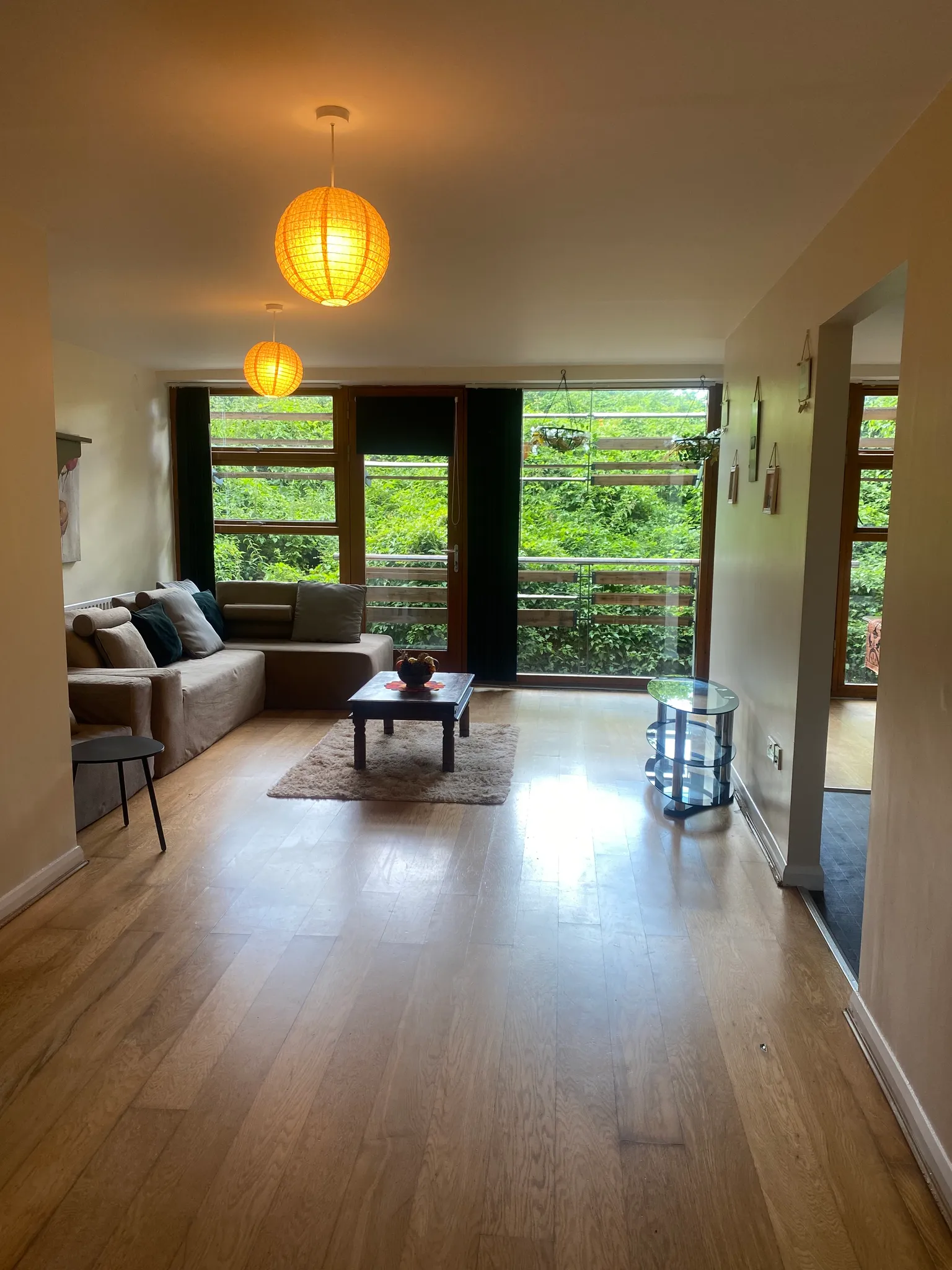 Living room with a sectional sofa, coffee table on a rug, pendant ceiling lights and large floor-to-ceiling windows/door opening to greenery.