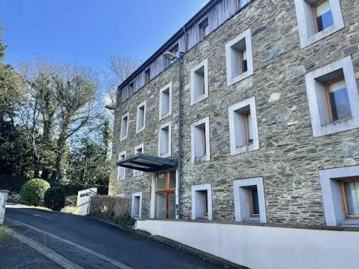 Exterior view of a multi-story stone apartment building with multiple rectangular windows, a recessed entrance with a small canopy, a paved driveway and trees to the left under a clear sky.