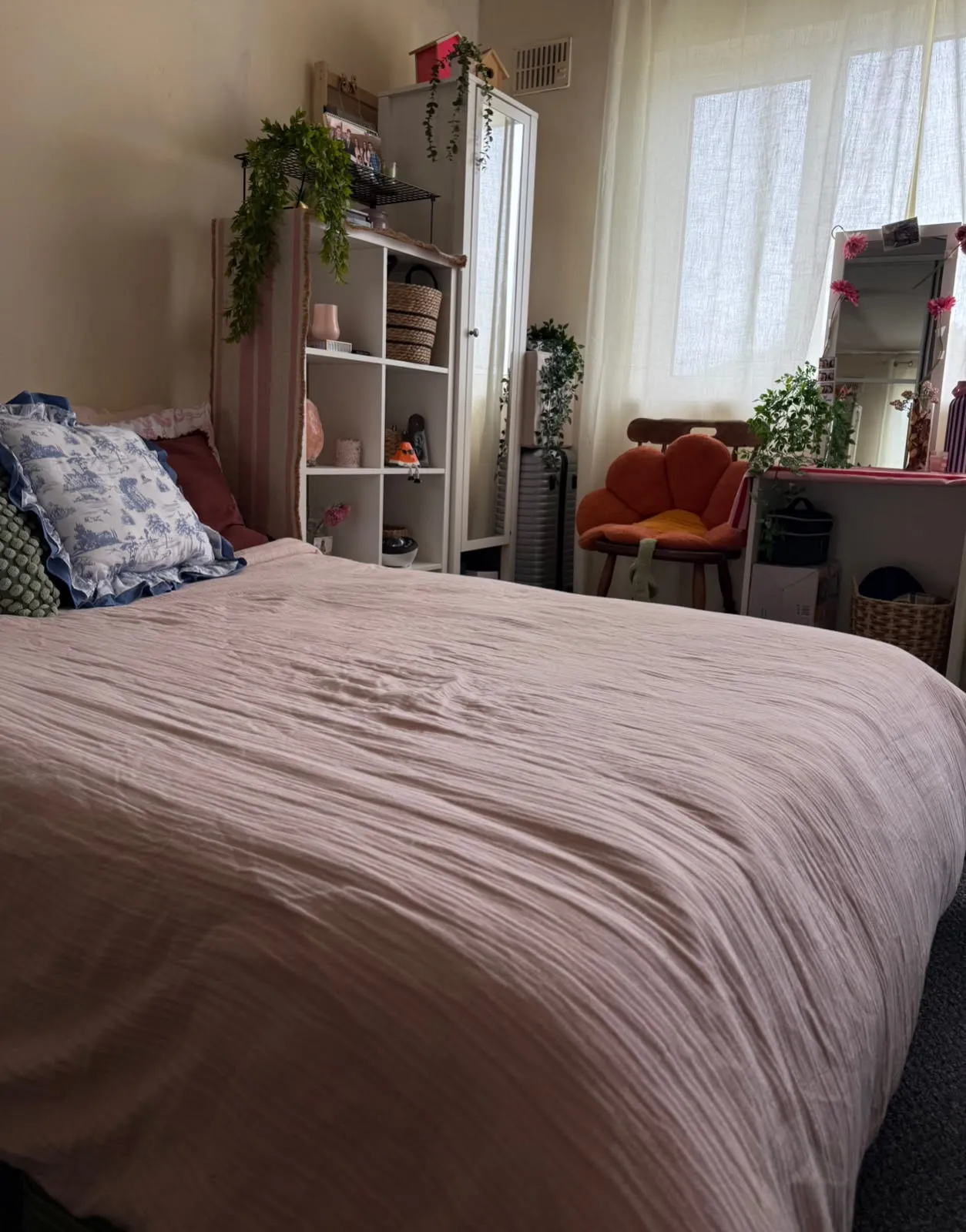 Bedroom with a large bed in the foreground, pillows on the left, a shelving unit and tall mirror against the wall, a window with sheer curtains, a small chair and plants near a vanity on the right.