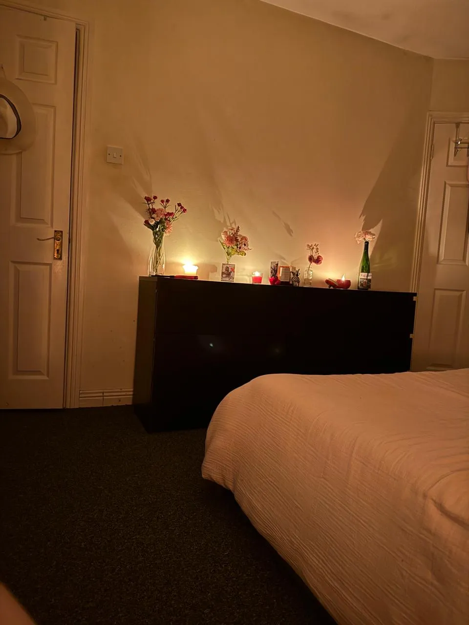 Bedroom with a bed in the foreground, a dark dresser against a wall topped with candles and vases of flowers, and two closed doors.