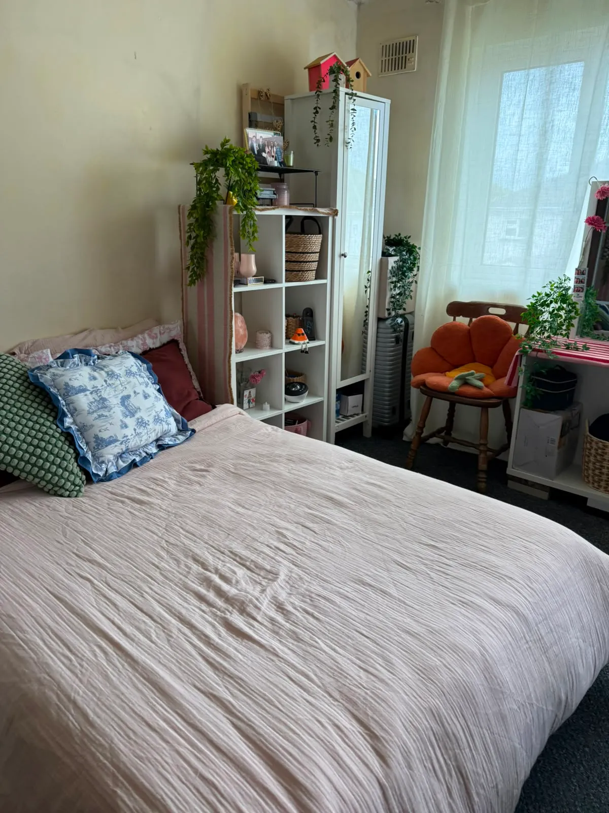Bedroom with a double bed, pillows, shelving unit with decorative items, a full-length mirror, chair, and a window with sheer curtains.