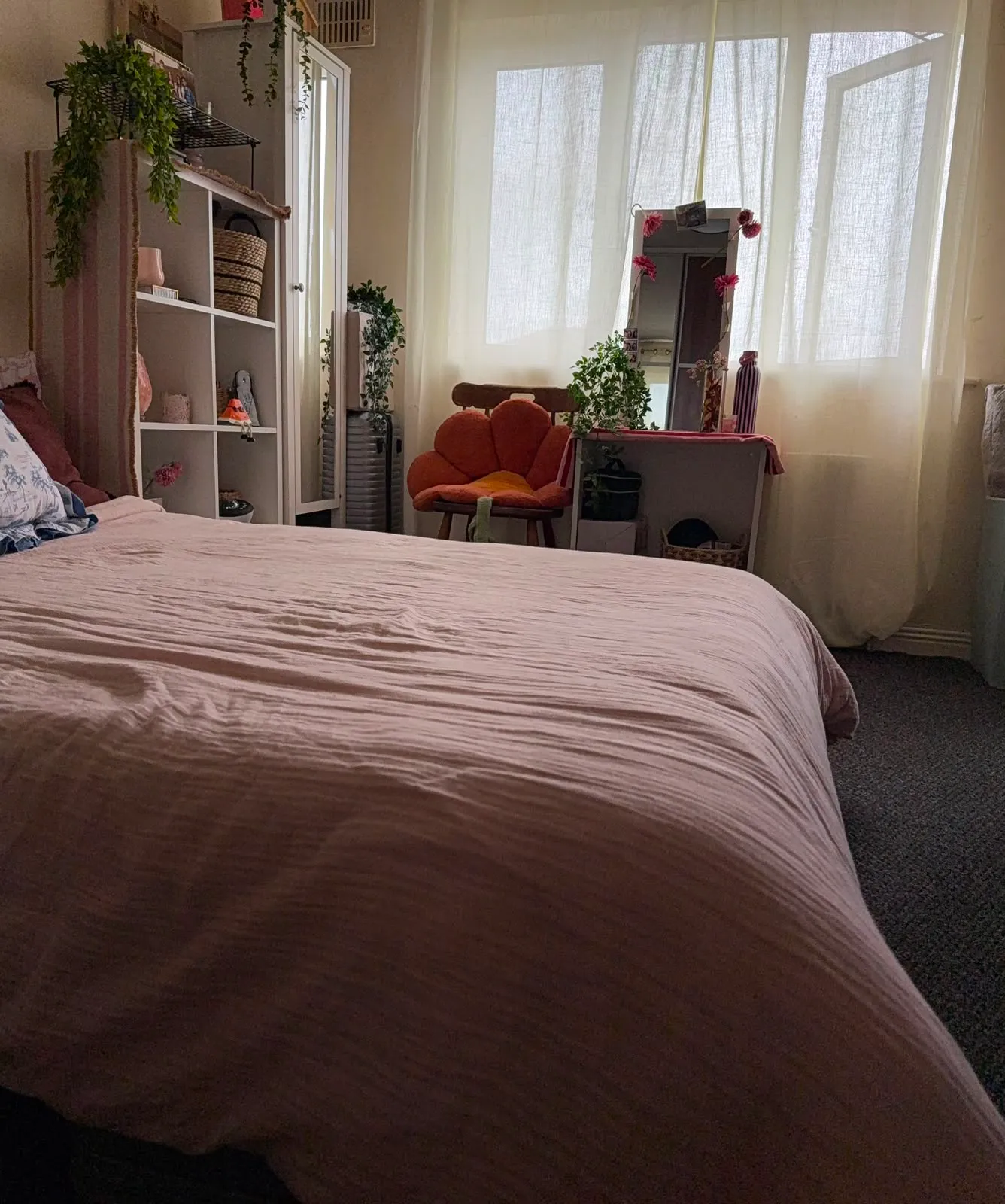 Bedroom with a large bed in the foreground, sheer-curtained window, a small desk or vanity with a mirror and plants, shelving unit and an orange upholstered chair.