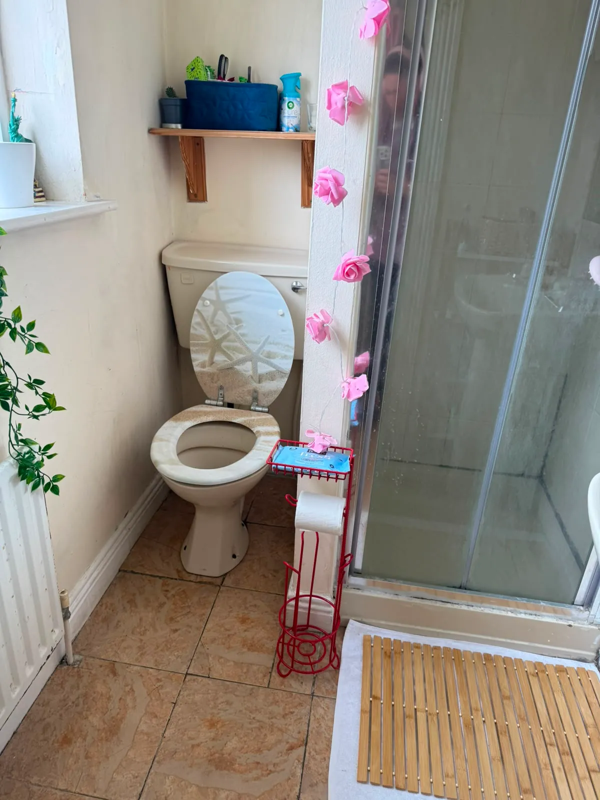 Bathroom with a toilet, shower with glass door, small shelf above the toilet, and a freestanding toilet paper holder.