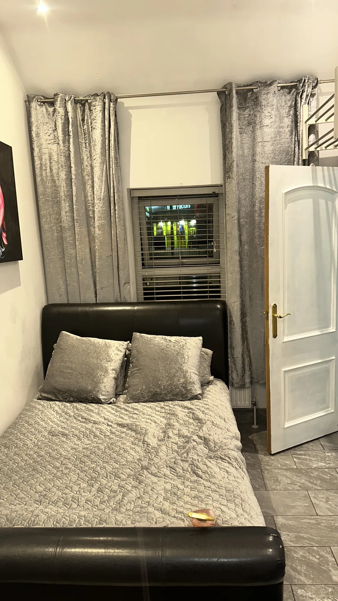 Bedroom with a double bed and headboard, two pillows, a small window with blinds and grey curtains, and a partially open white door.