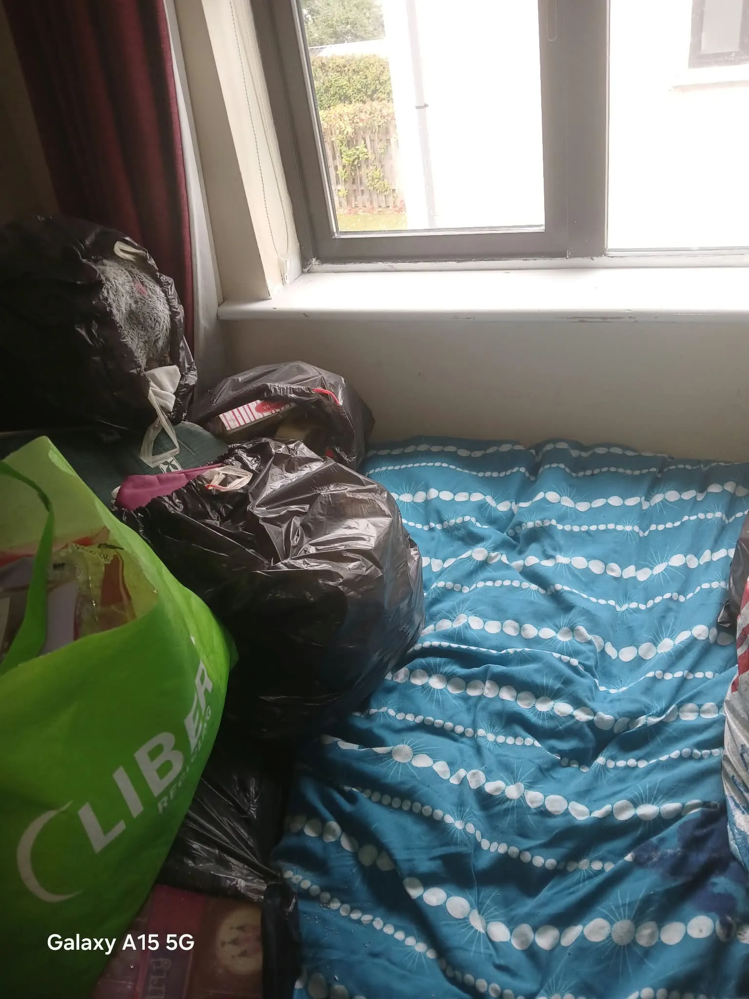 Bedroom window area with a mattress or bedding on the floor, blue patterned duvet, several black and green bags near the window and a partially visible curtain.