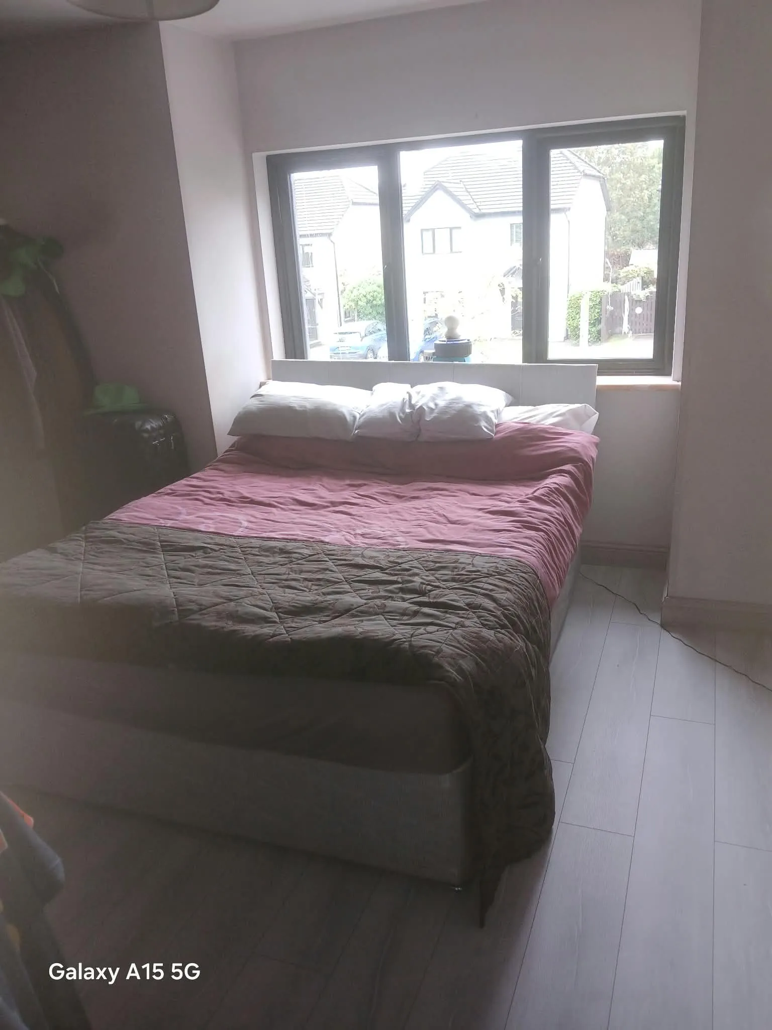 Bedroom with a double bed positioned under a wide three-panel window, pillows on the headboard and a visible suitcase at left.