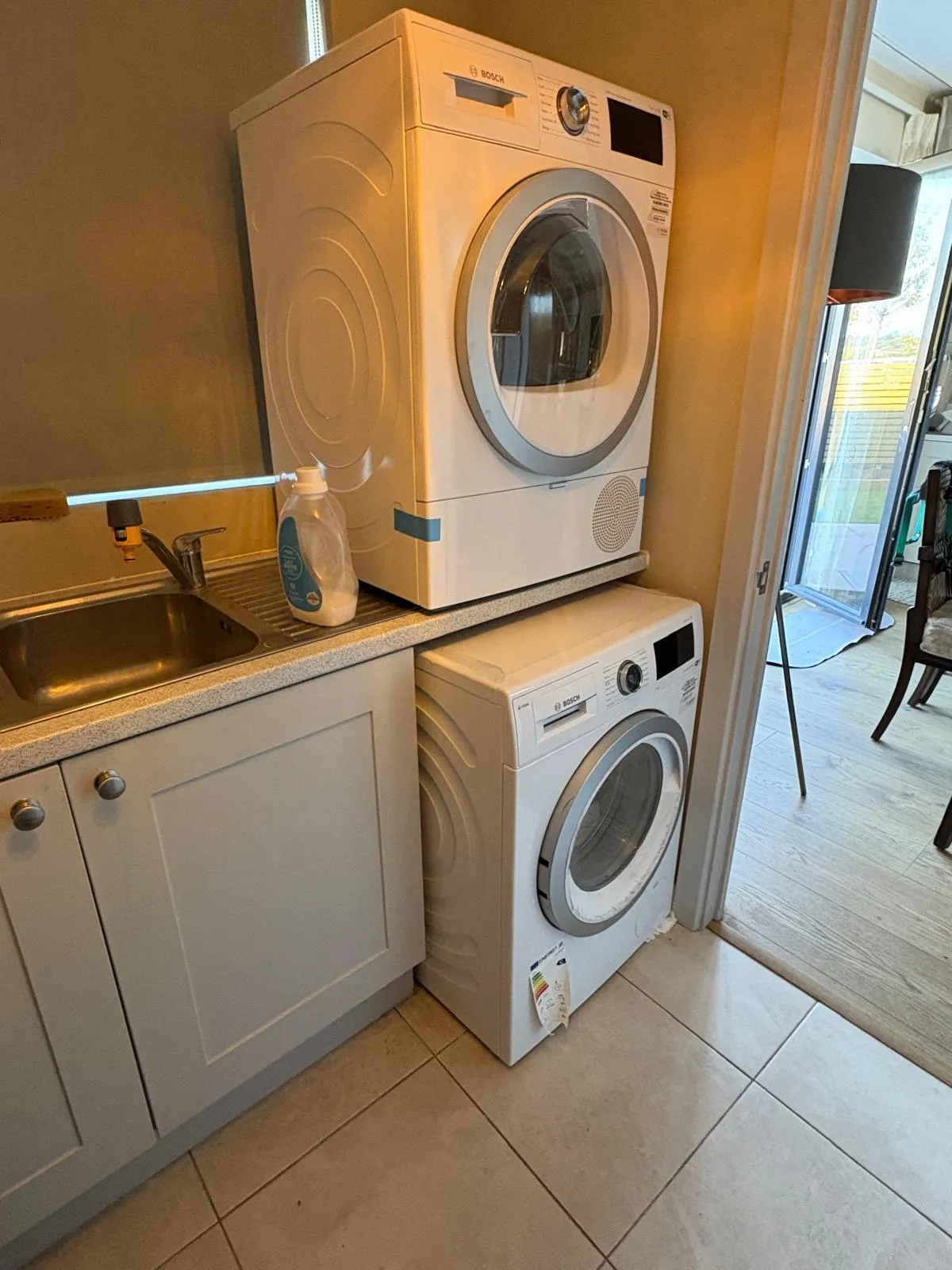 Utility area with stacked front-load washer and dryer next to a sink and countertop, tiled floor and open doorway.