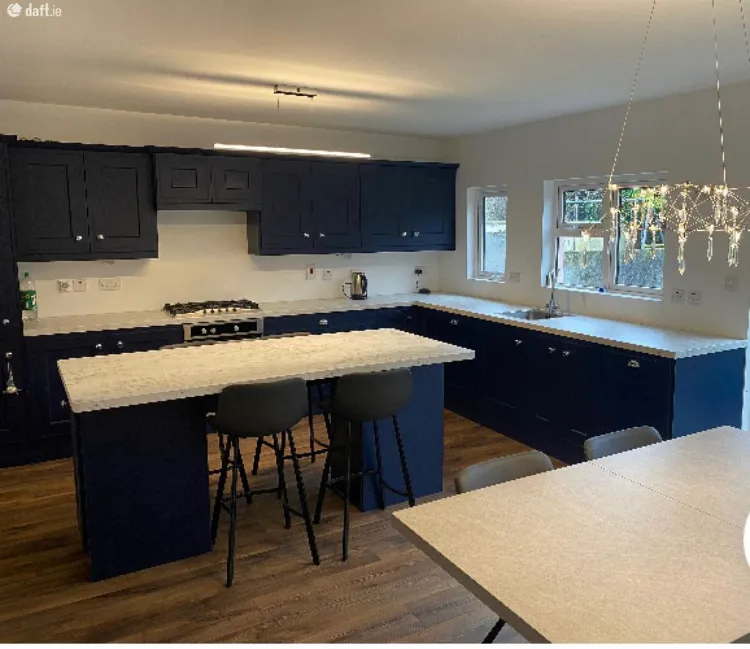 Kitchen with a central island, two bar stools, dark blue lower and upper cabinets, marble-effect countertops, gas stove, sink under two windows, and a hanging light fixture