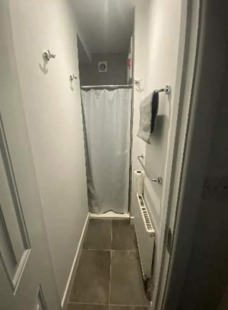 Narrow bathroom corridor with tiled floor, radiator and towel rack on the right, wall hooks on the left, and a shower with a closed curtain at the far end.
