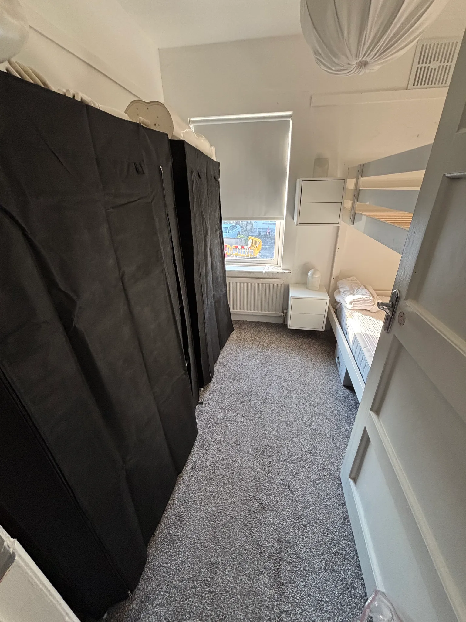 Small bedroom with a bunk bed on the right, fabric-covered wardrobe on the left, a window with a roller blind, small bedside table and radiator.