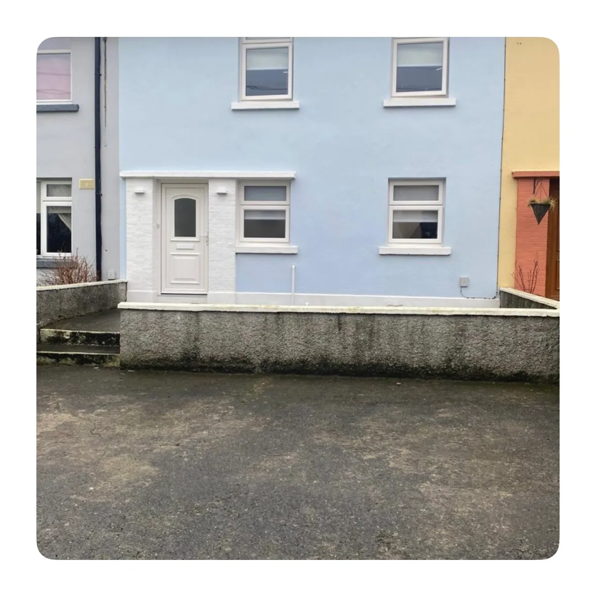 Front exterior of a two-story pale blue house with a white entrance door, three ground and first-floor windows, a low concrete wall and paved area in front.