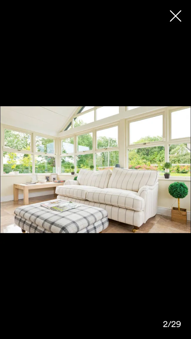 Sunroom with large windows overlooking garden, cream striped sofa, checkered ottoman, wooden side table, and potted plant
