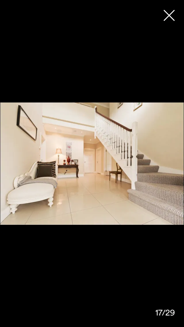 Hallway with tile flooring, white upholstered chair on left, staircase with striped carpet runner on right, framed artwork on wall, and doorways leading to other rooms