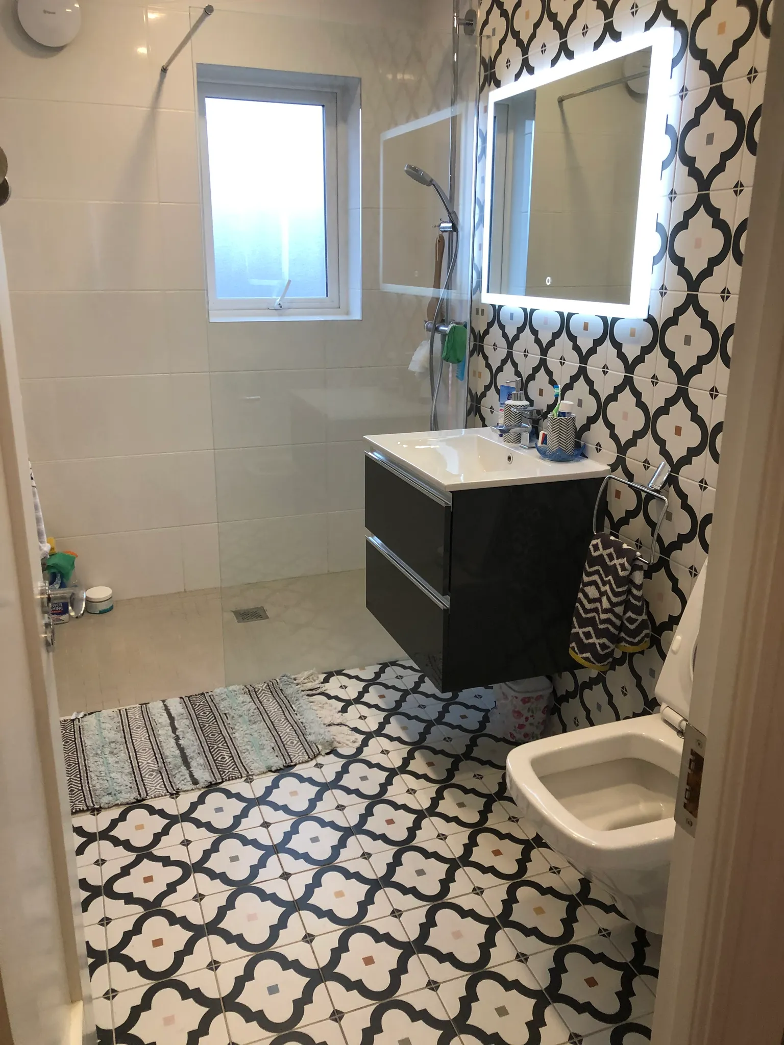 Bathroom with patterned floor and wall tiles, floating vanity with sink, illuminated mirror, shower area with glass screen and frosted window, and a wall-mounted toilet partially visible.