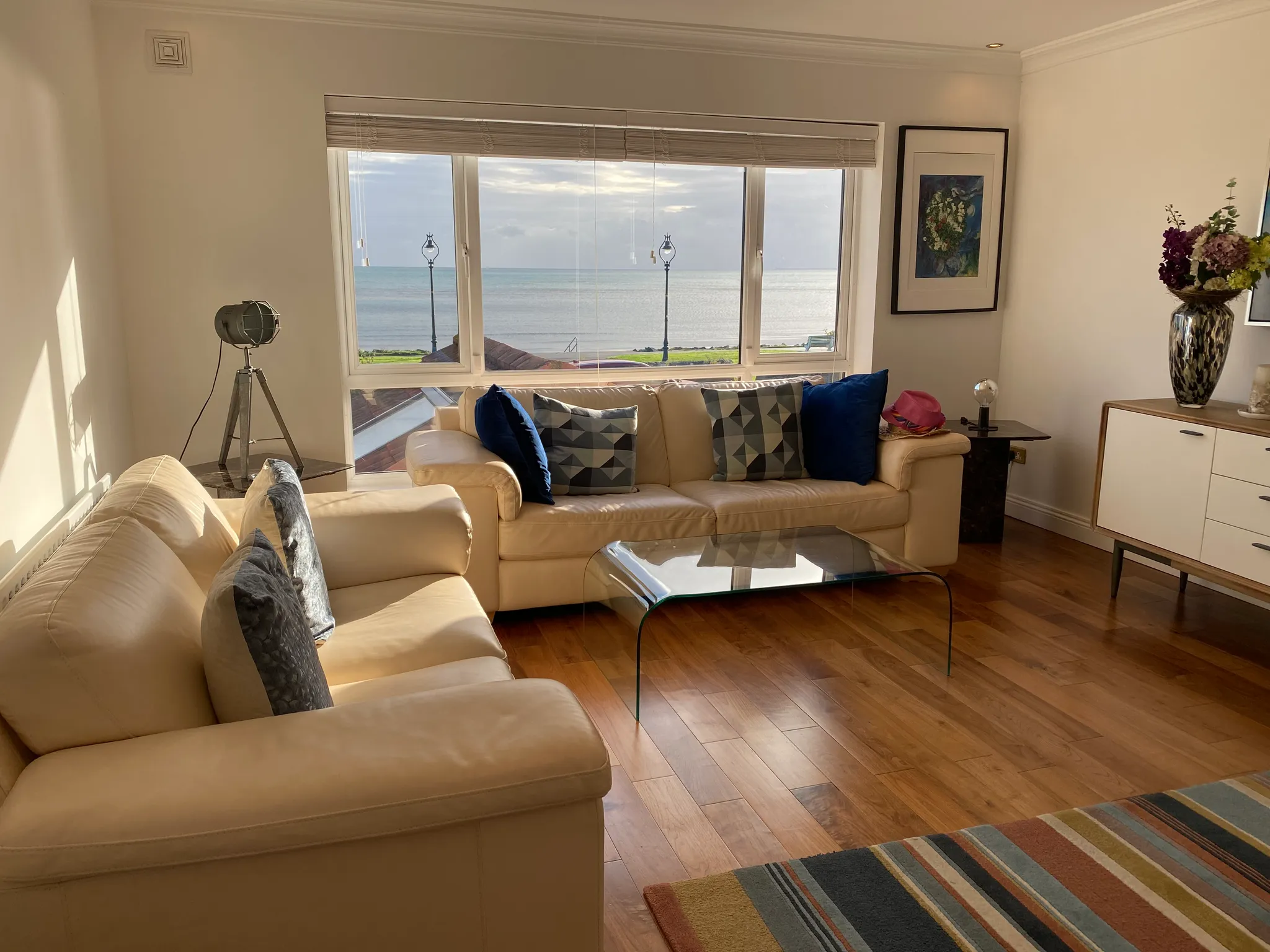 Living room with beige sofas, patterned cushions, glass coffee table, large window with sea view and blinds, floor lamp and sideboard with vase of flowers.
