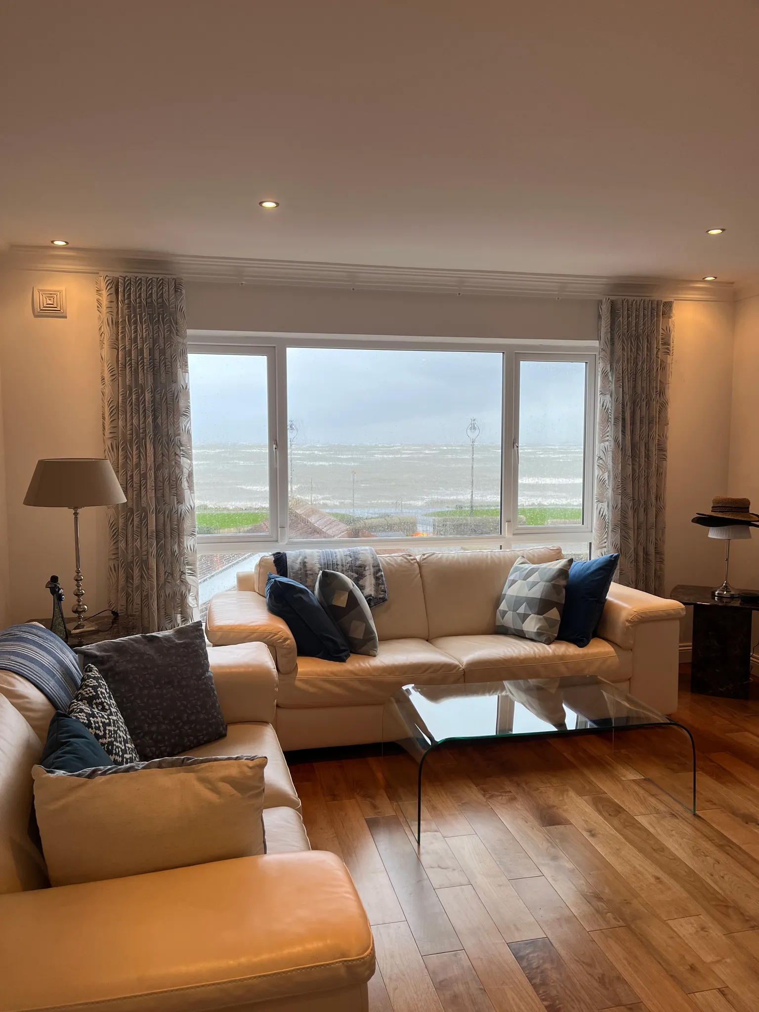 Living room with cream leather sofas, patterned cushions, a glass coffee table and a large window with curtains showing a seascape outside.