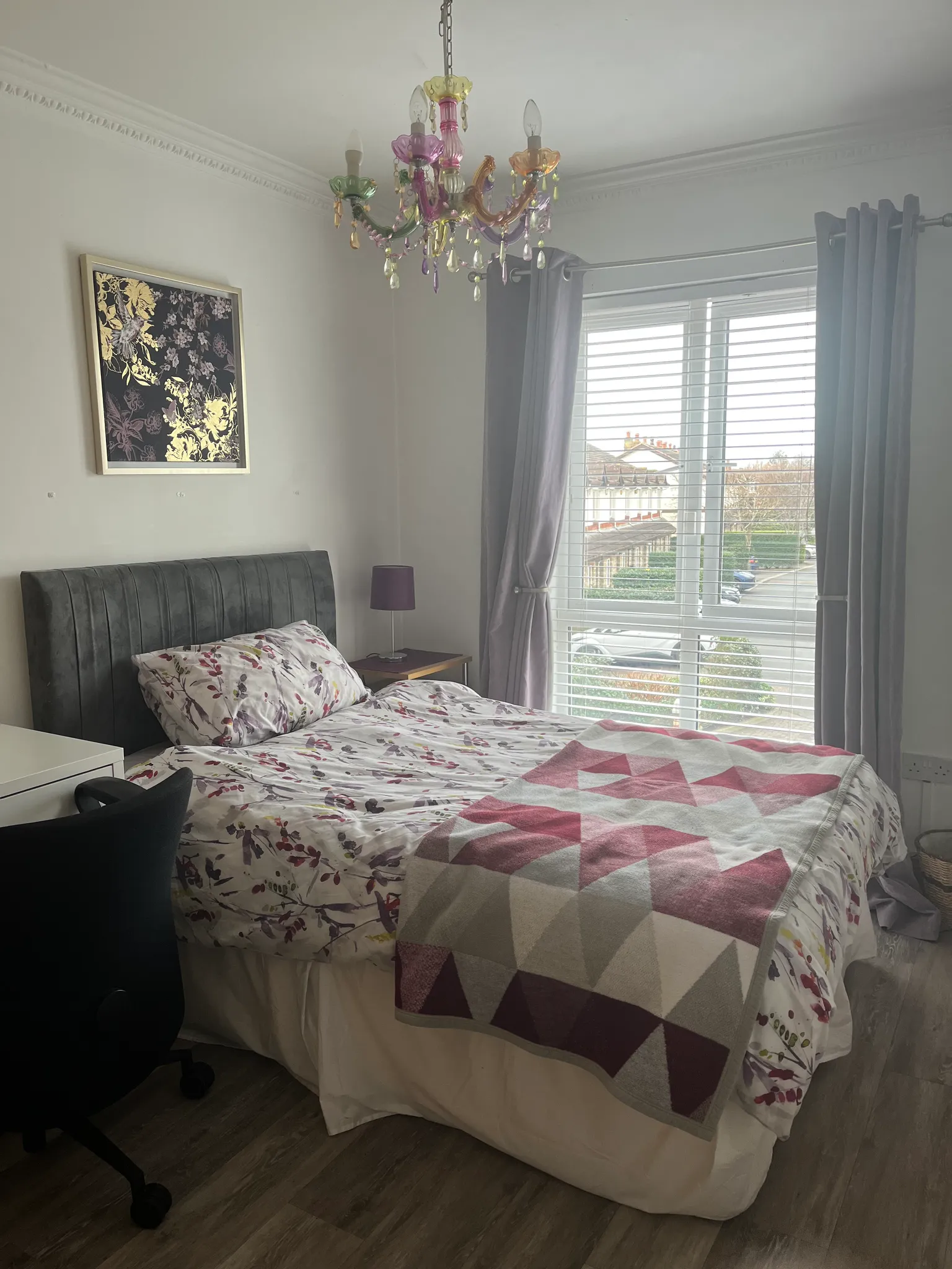 Bedroom with a double bed, patterned bedding, upholstered headboard, bedside table with lamp, window with blinds and curtains, framed wall art and a colorful ceiling chandelier.