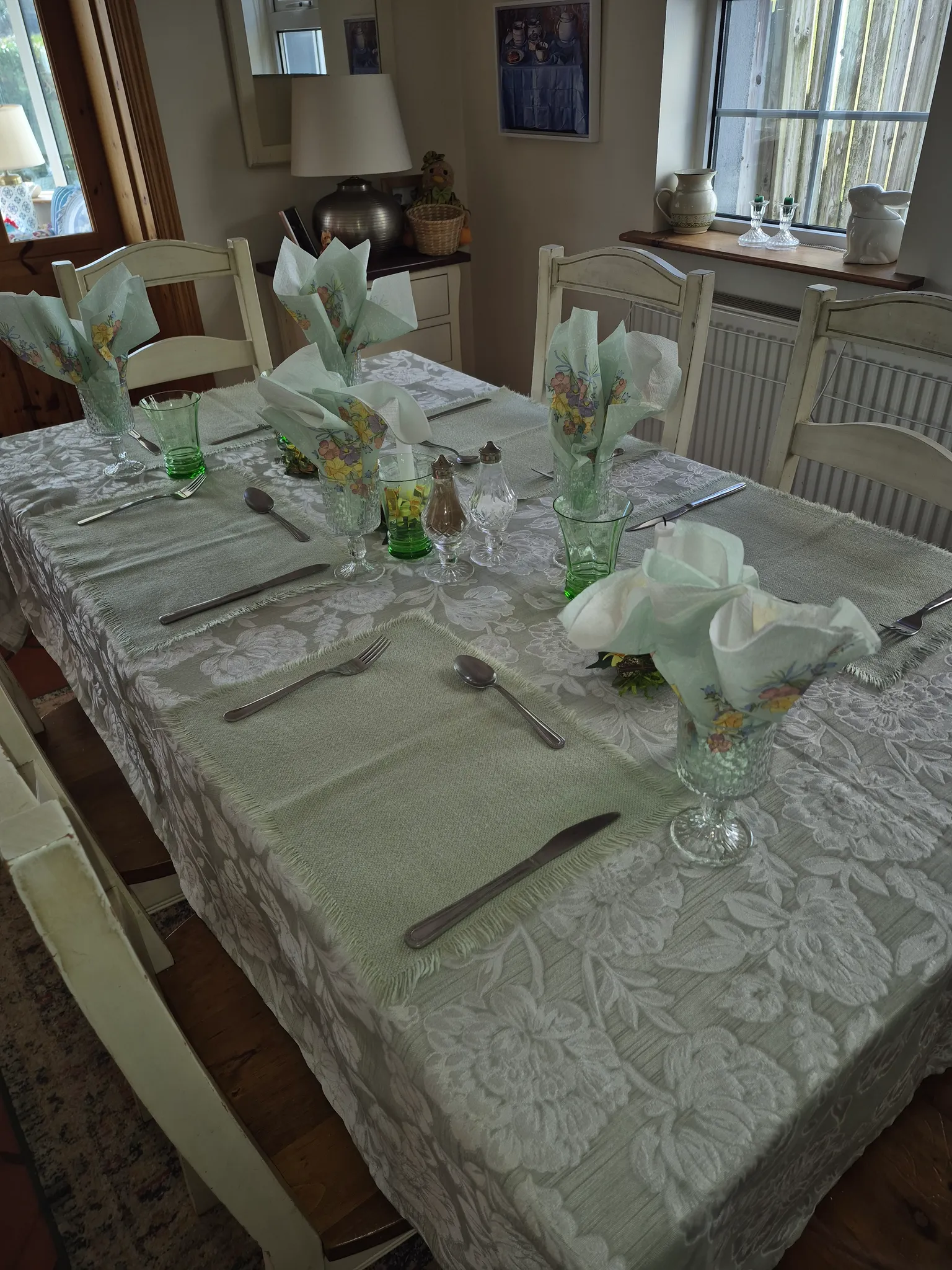 Dining room with a rectangular table set for a meal, folded floral napkins in glassware, cutlery and green glasses, white wooden chairs and a window with a windowsill display.