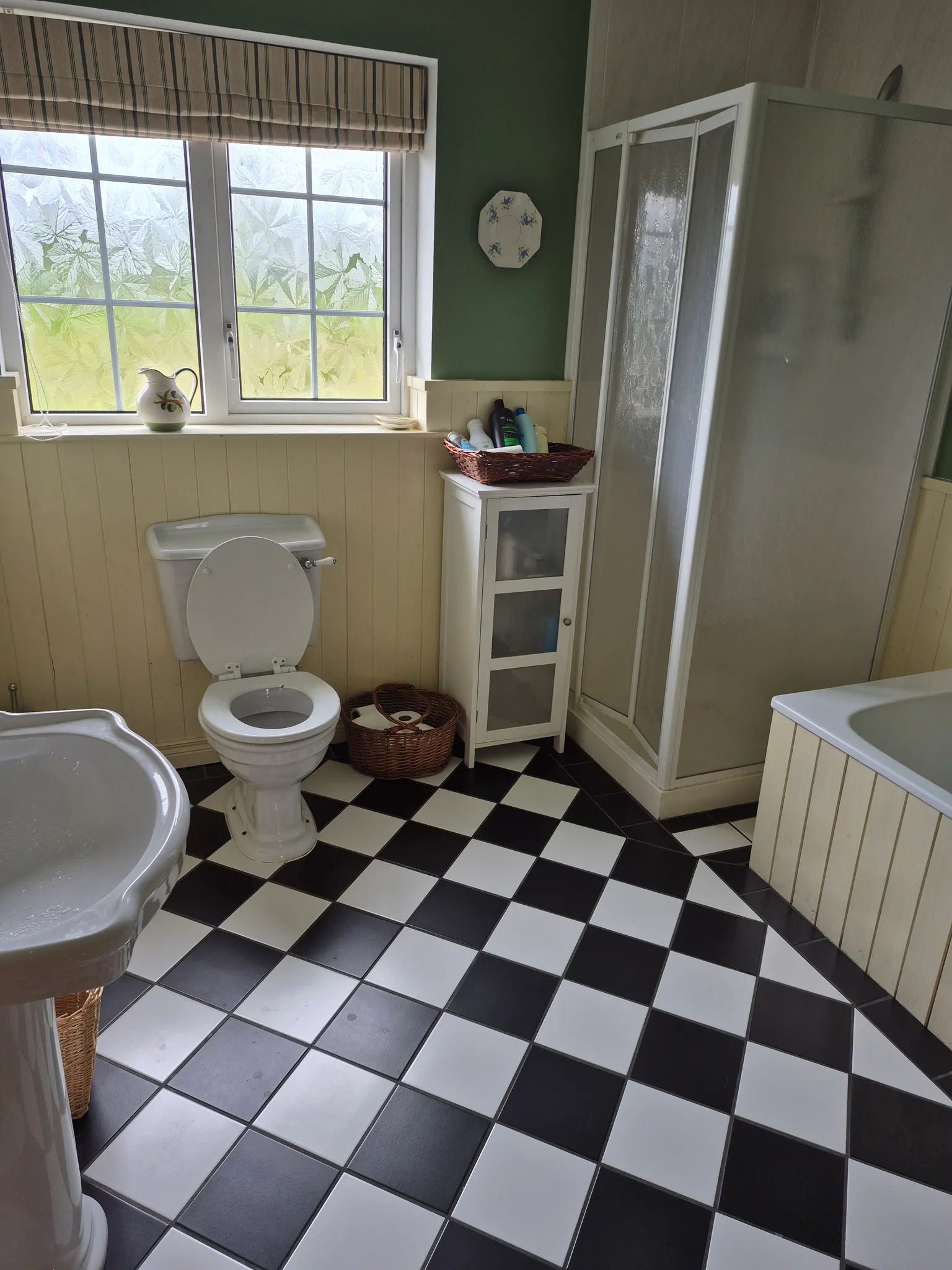 Bathroom with a toilet, pedestal sink, shower cubicle, bathtub edge, frosted window with blinds and a black-and-white checkered tile floor.