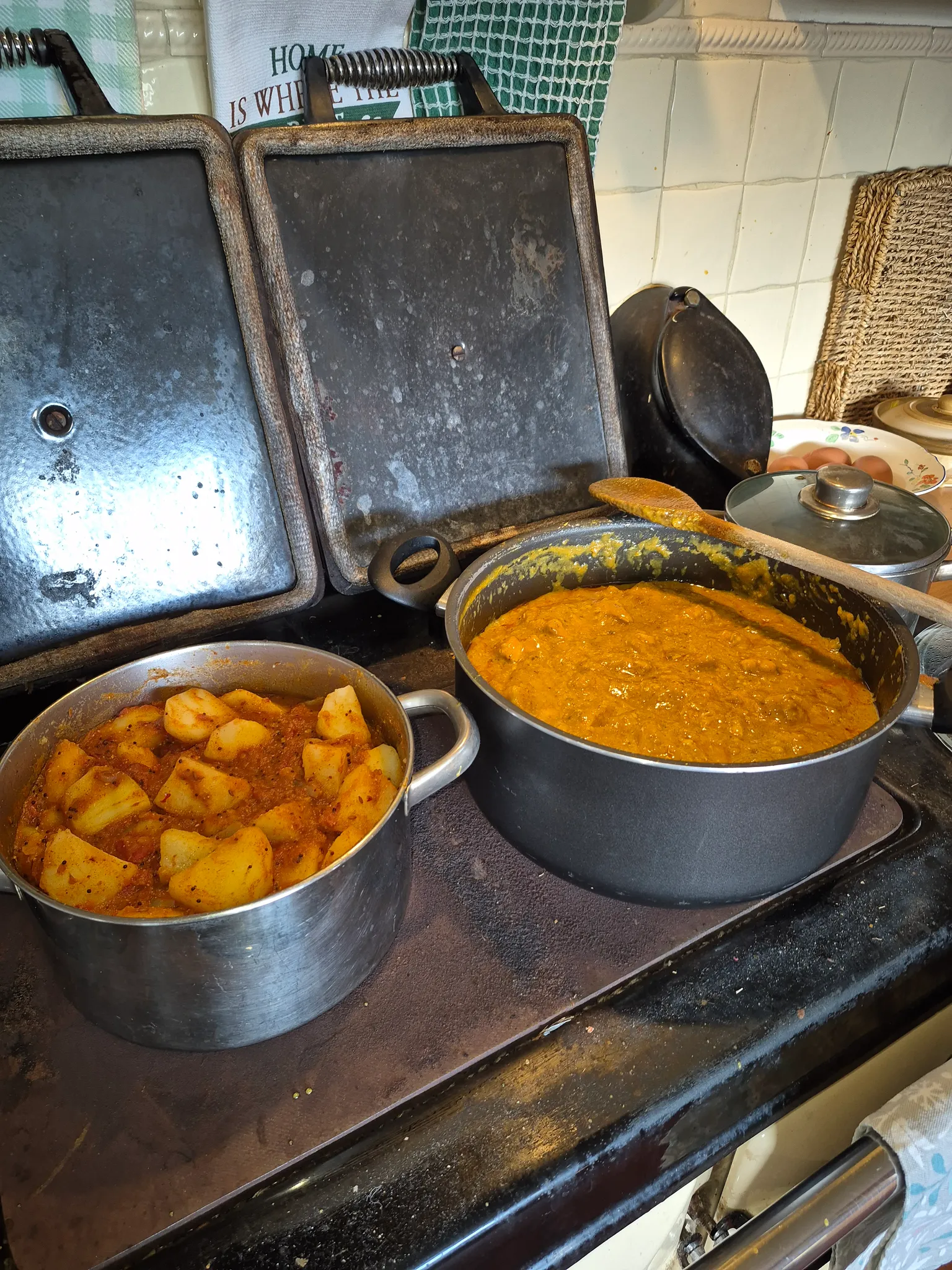 Kitchen stovetop with two pots of cooked food, a wooden spoon, and two rectangular griddle plates propped against a tiled backsplash.