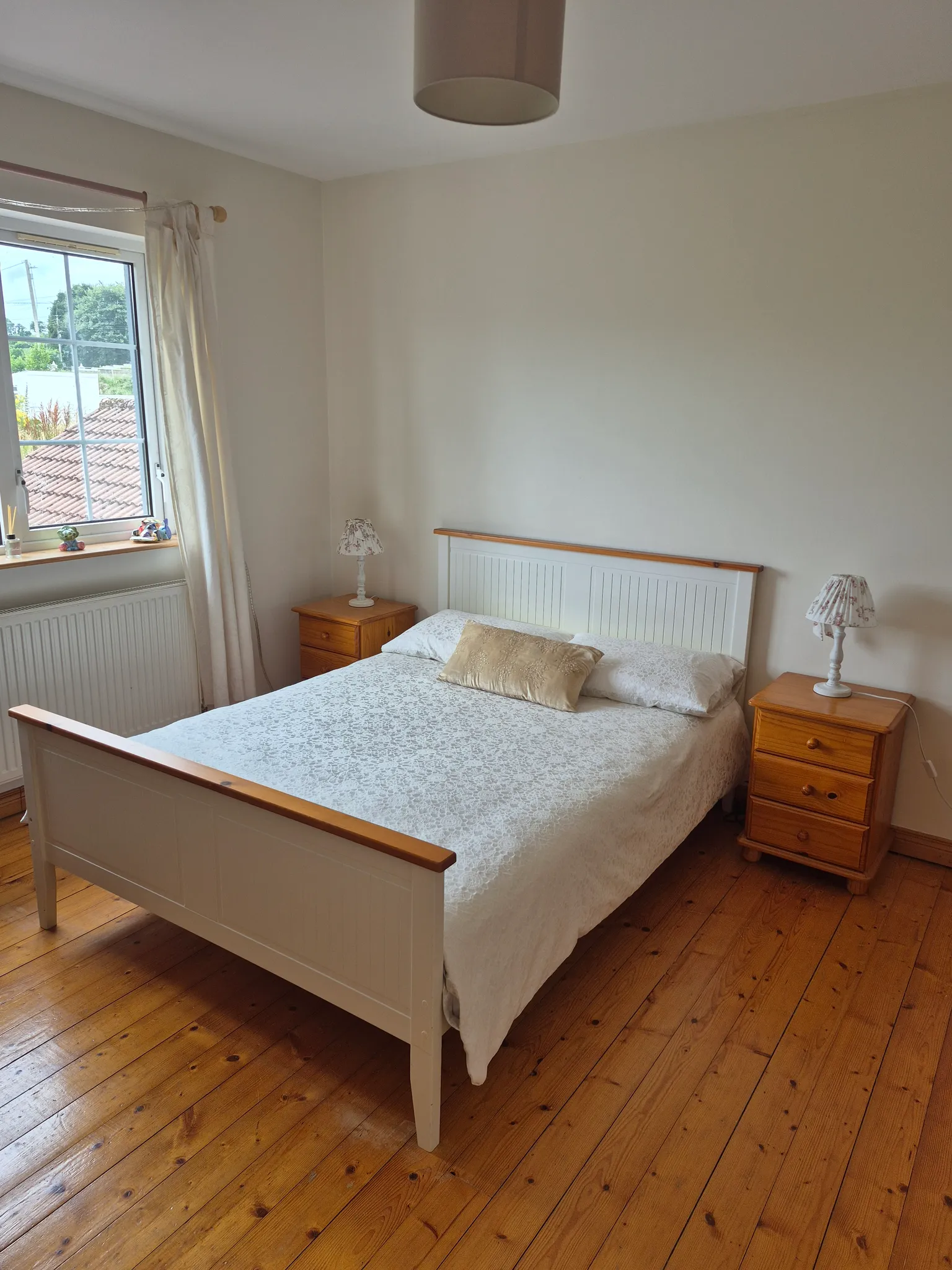 Bedroom with a double bed and headboard, two wooden nightstands with lamps, a window with curtains, and wooden floor.