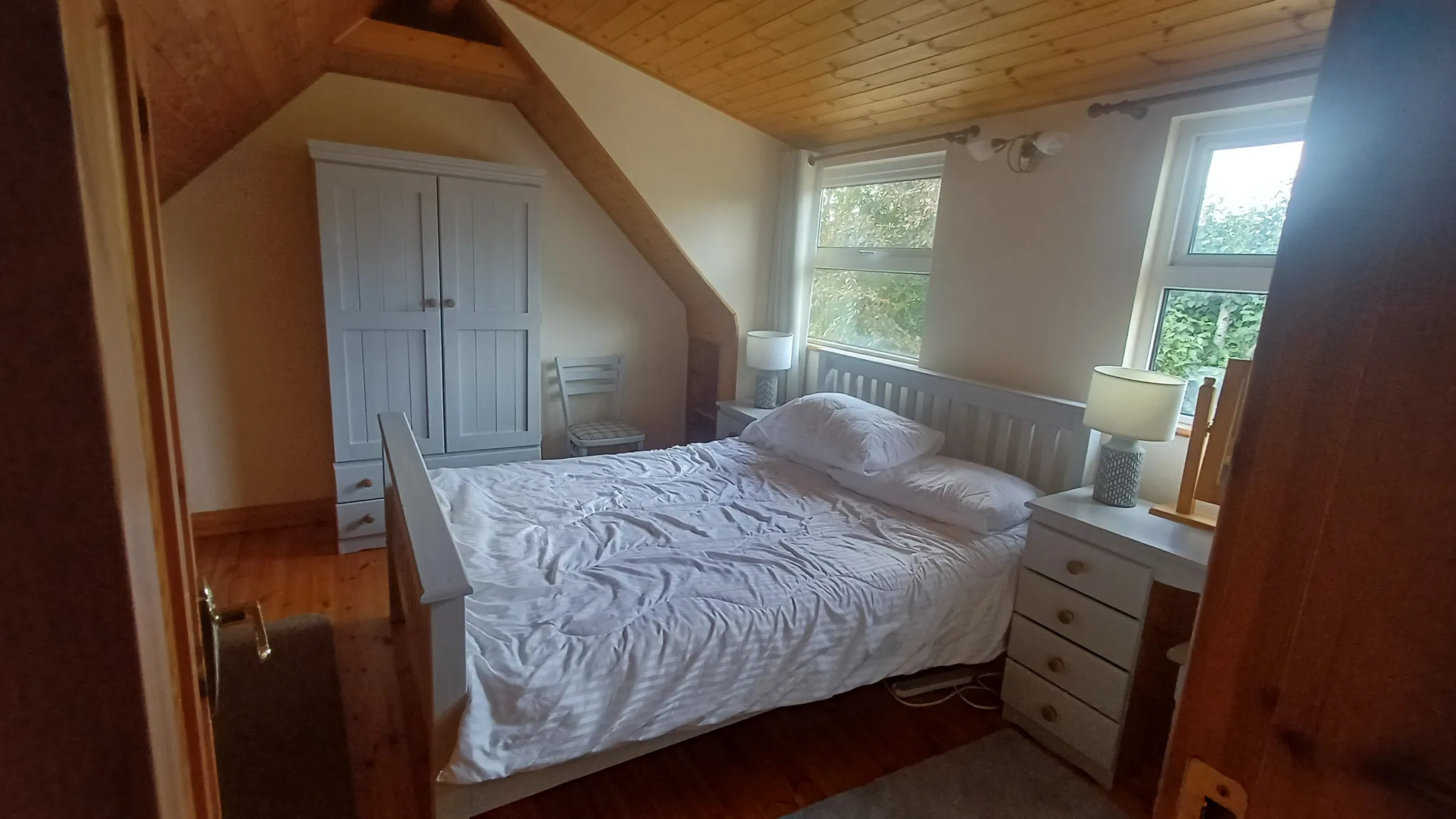 Bedroom with a double bed, two bedside tables with lamps, a white wardrobe, a chair, and two windows under a sloped wooden ceiling.