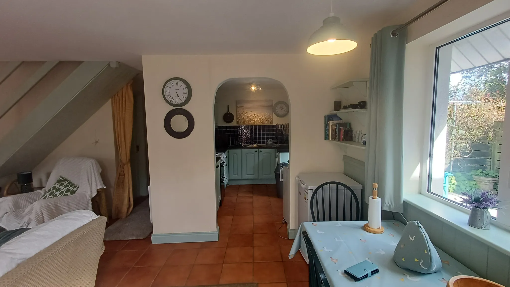 Open-plan living area with a sofa and armchair on the left, a dining table by a large window on the right, a pendant light overhead and an arched doorway leading to a small kitchen.