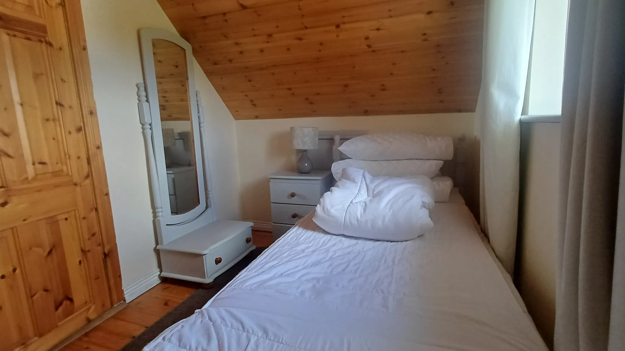 Bedroom with a single bed, stacked pillows and duvet, bedside table with lamp, free-standing mirror, slanted wooden ceiling and a window with curtains.