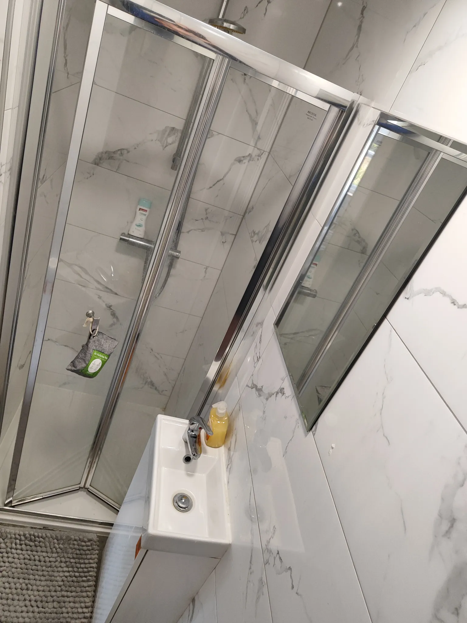 Bathroom with a glass-enclosed corner shower, visible showerhead and toiletries, a small sink with faucet and soap bottle, and a wall-mounted mirror.