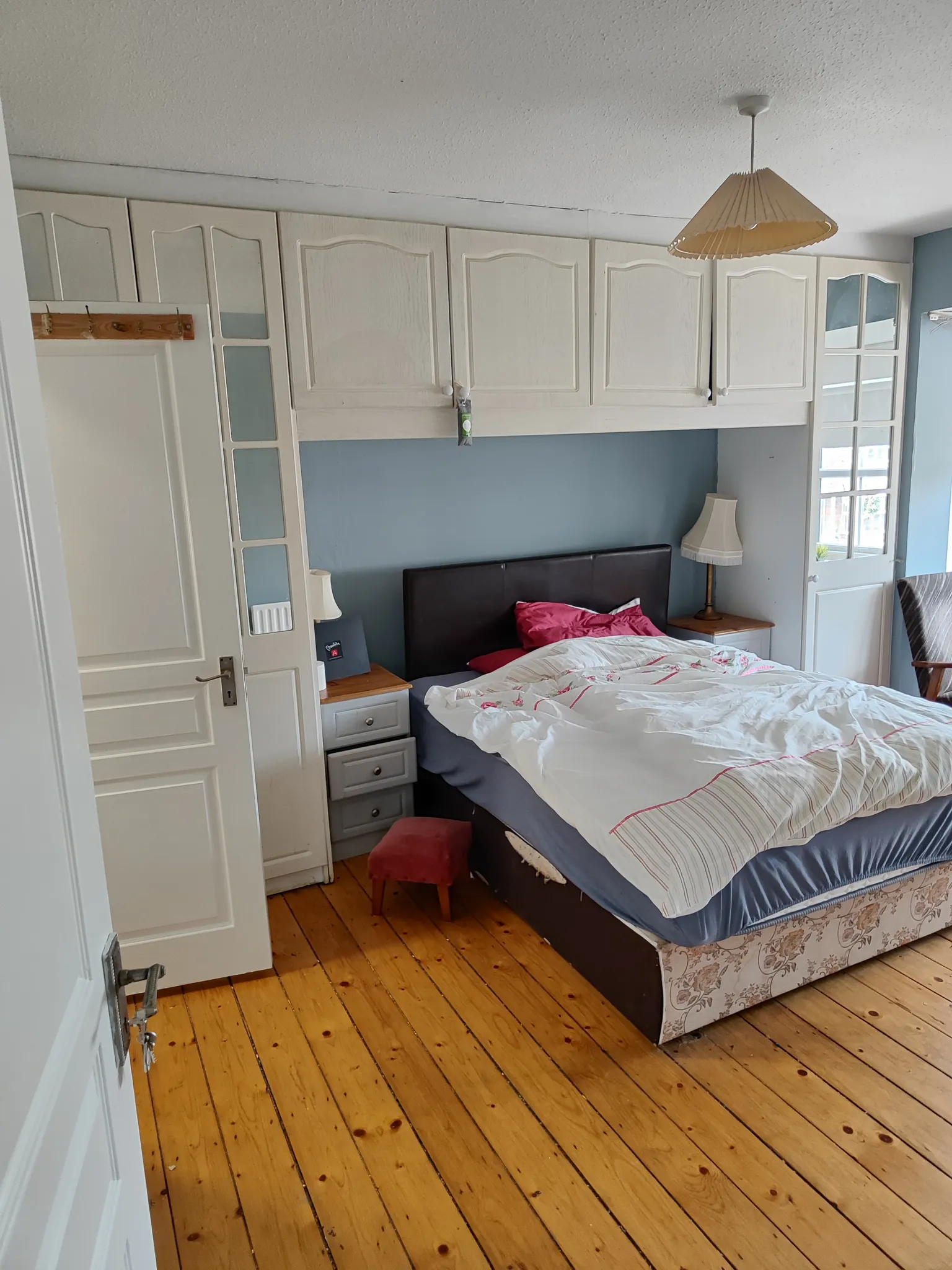 Bedroom with a double bed, overhead white cabinets, bedside tables with lamps, wooden floor and a pendant ceiling light