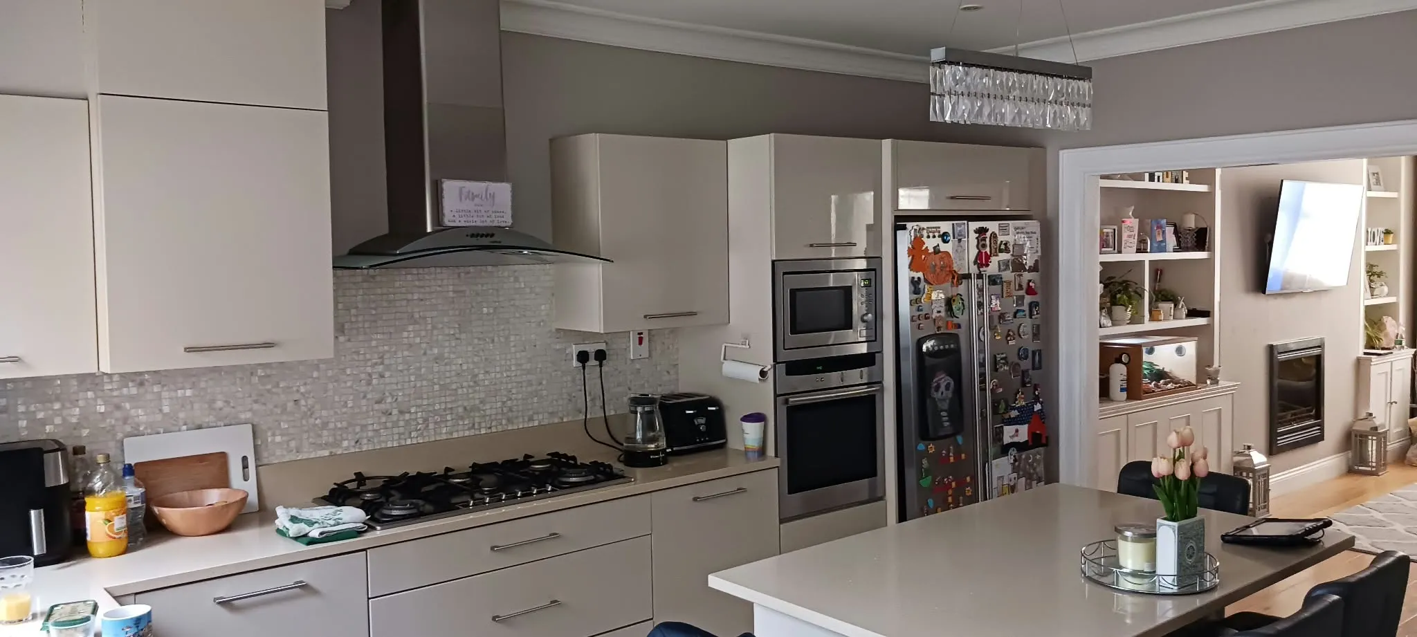 Kitchen with countertop island, built-in oven and microwave, gas cooktop with range hood, refrigerator covered in magnets, cabinets and open shelving visible into an adjacent living area.