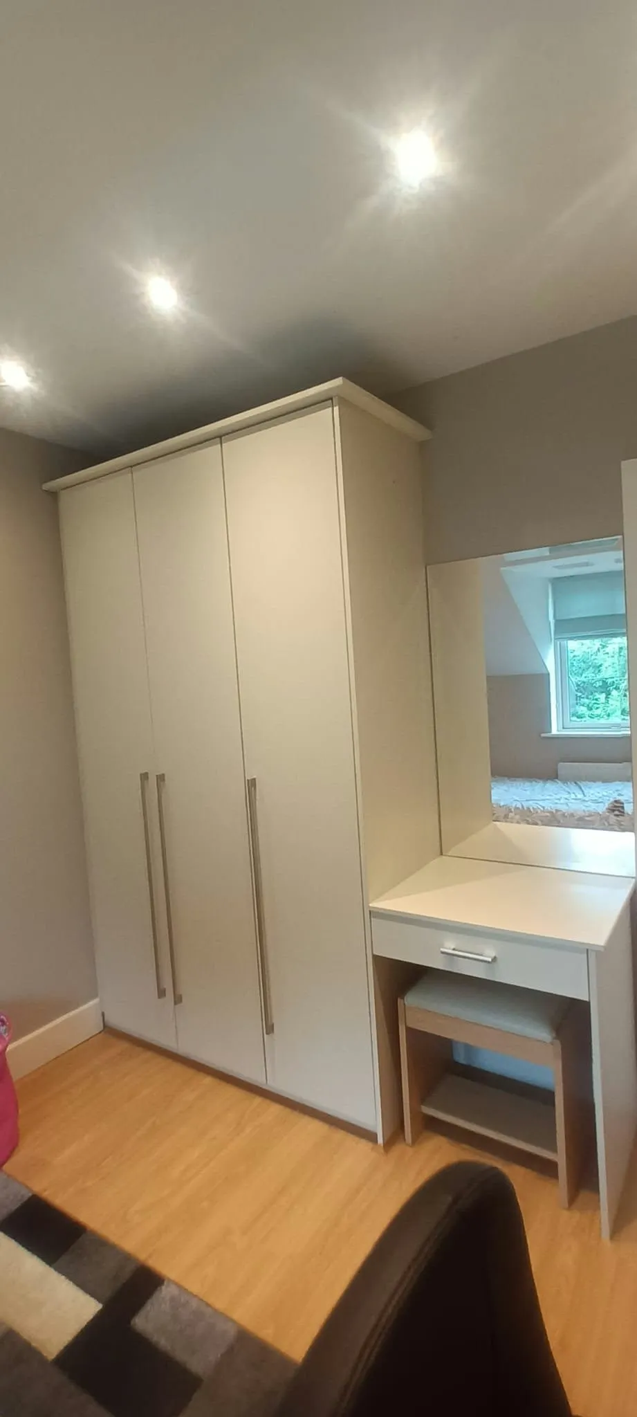Bedroom corner with a tall white wardrobe, a small white vanity desk with mirror and stool, hardwood floor and recessed ceiling lights.
