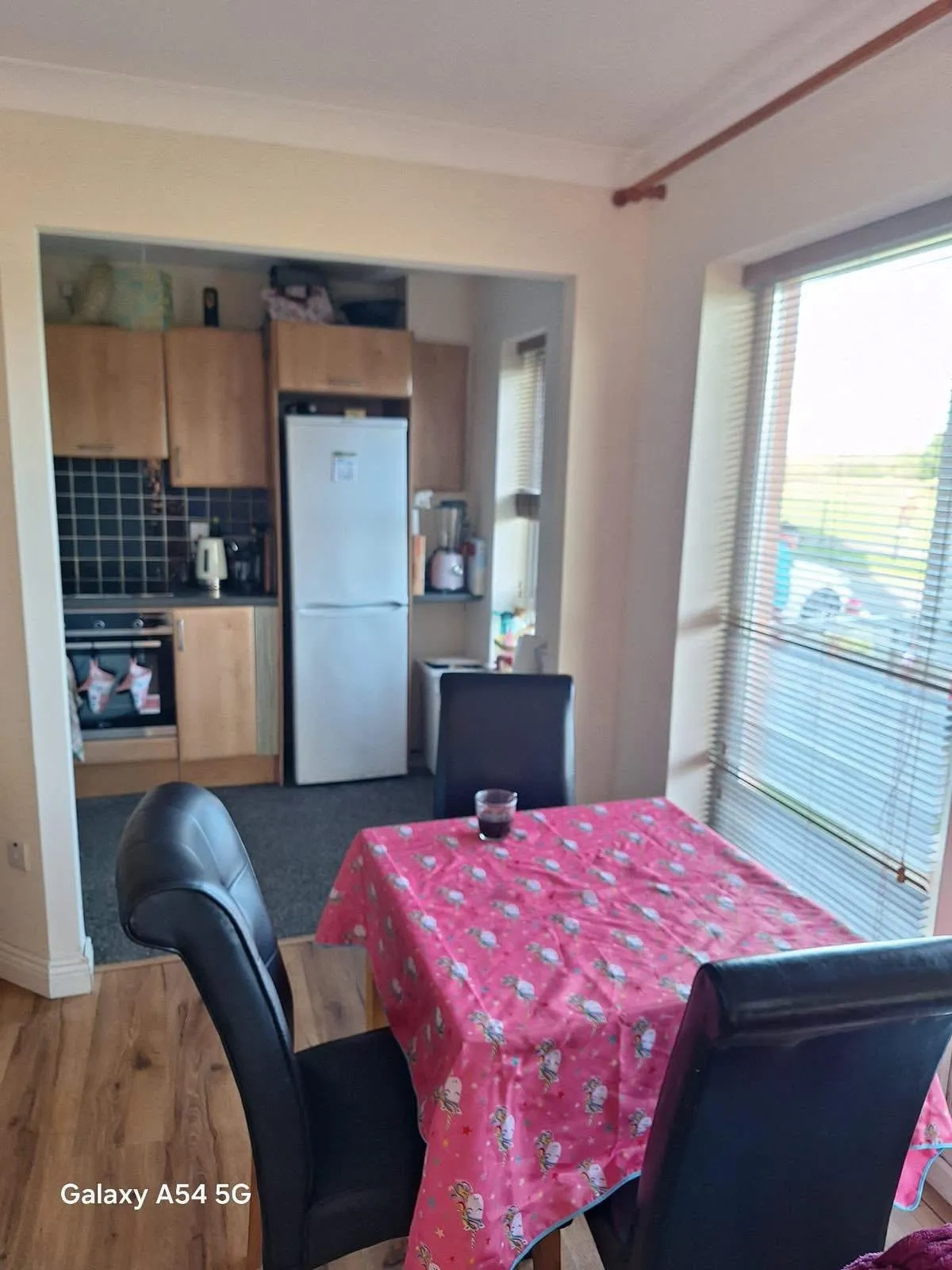 Dining area with a pink tablecloth on a rectangular table, several black dining chairs, a large window with blinds, and an open view into a small kitchen with a refrigerator and wooden cabinets.