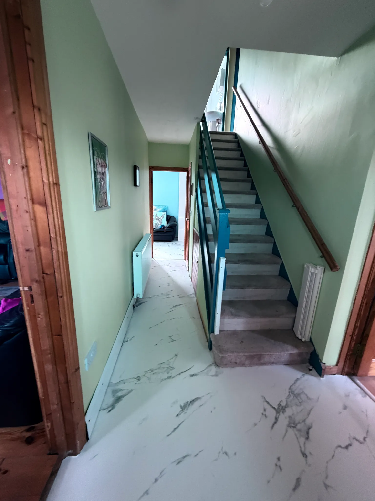 Hallway with a staircase on the right, wooden handrail, radiator on the left wall and a doorway leading to another room.