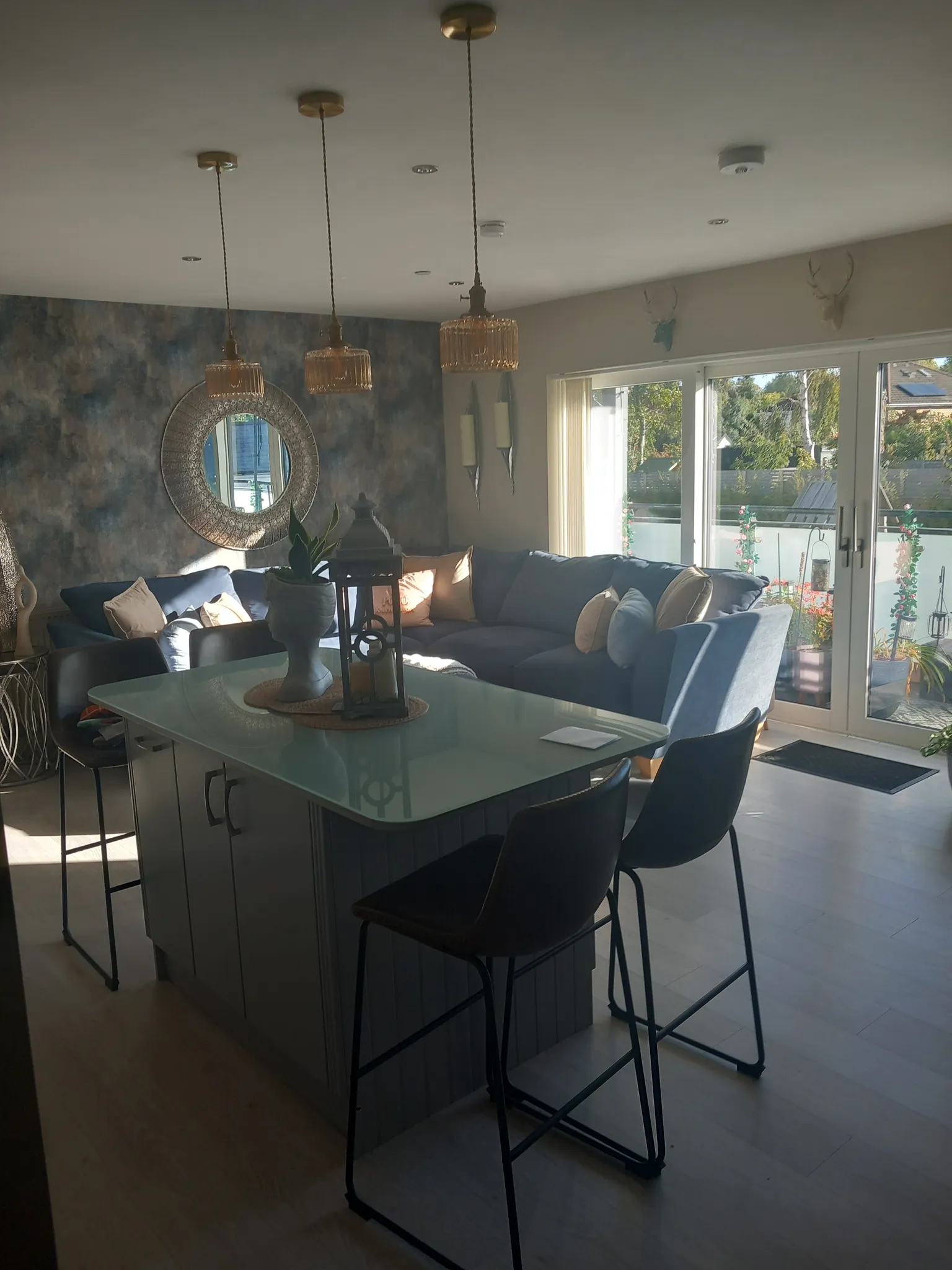 Living room with an island and bar stools in the foreground, sectional sofa with cushions, pendant lights, a round decorative mirror on a patterned wall, and sliding glass doors to the outside.