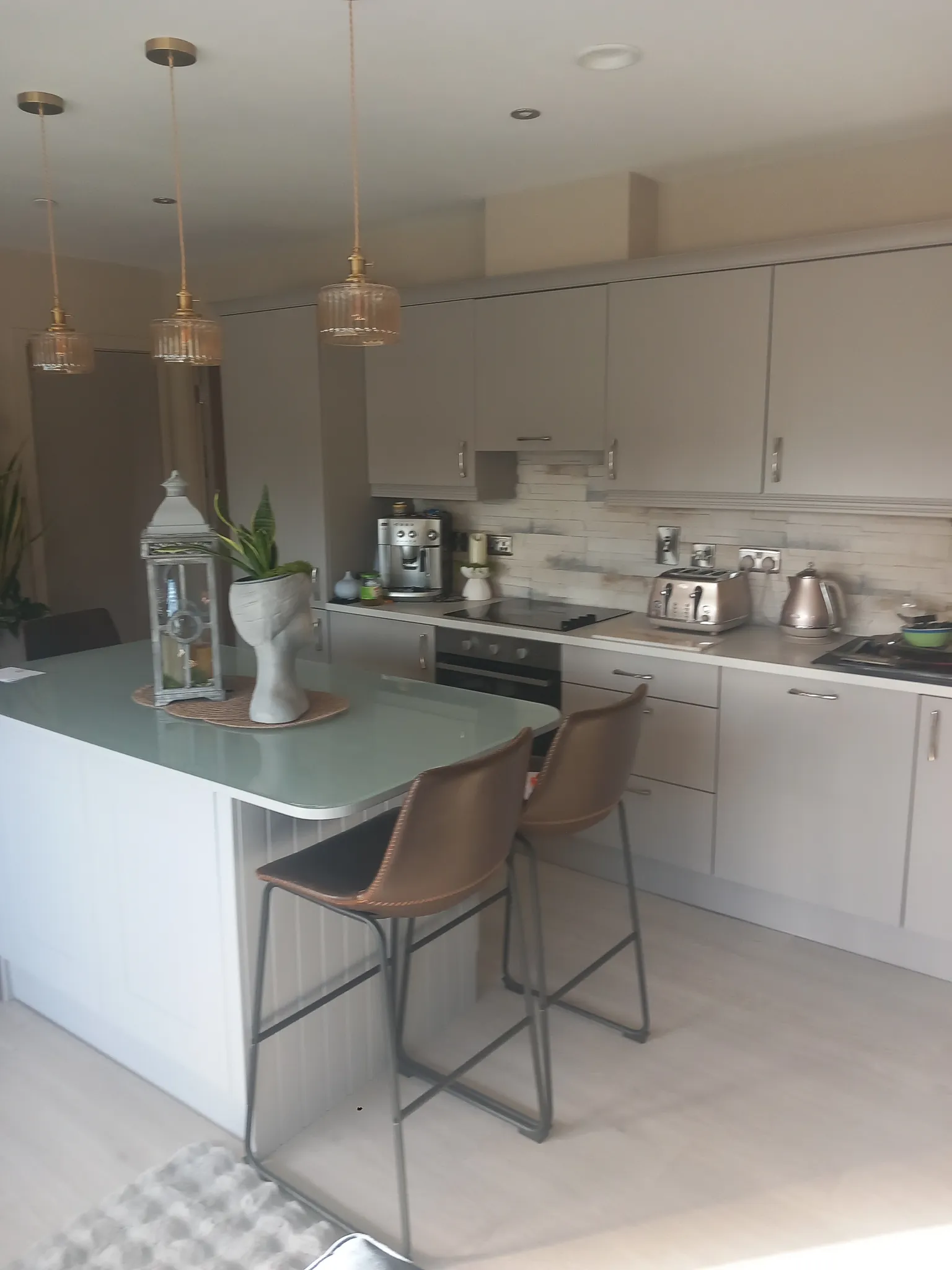 Kitchen with an island and two bar stools, overhead pendant lights, cabinets, countertop appliances (toaster, kettle, coffee maker) and a decorative plant.