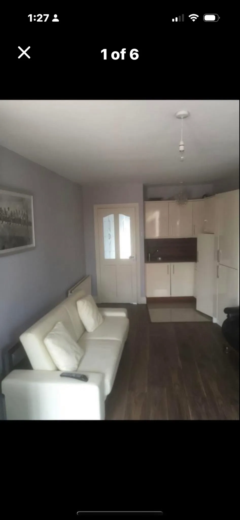 Living room with a white two-seat sofa and pillows, hardwood floor, small kitchenette with refrigerator and cabinets, and a closed interior door.